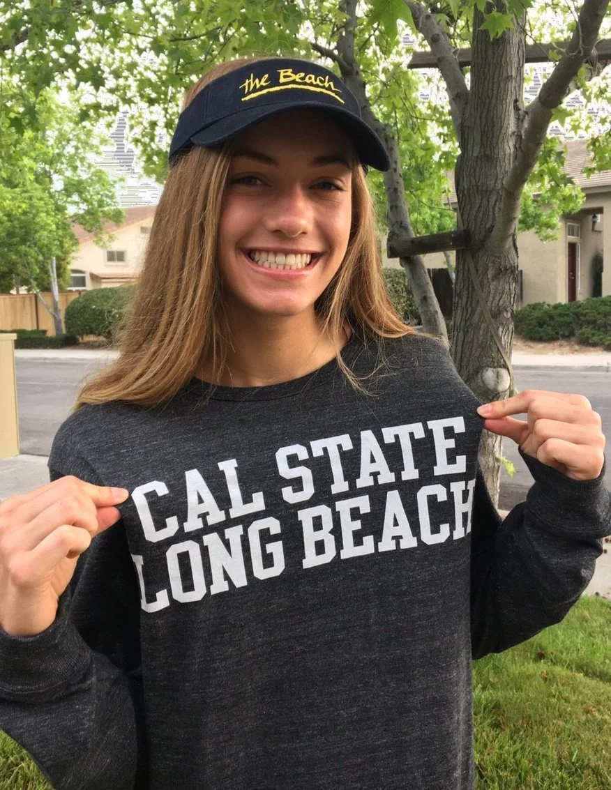 A young woman smiling outdoors, pointing to her sweatshirt that says 'Cal State Long Beach,' wearing a black visor with yellow writing that says 'The Beach.'