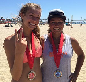 Two women at the beach wearing medals around their necks, smiling, with clear blue sky and beach scene in the background.