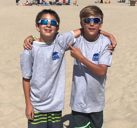 Two boys wearing gray T-shirts with a blue logo, sunglasses, and shorts, standing on a sandy beach with other people and beach equipment in the background.