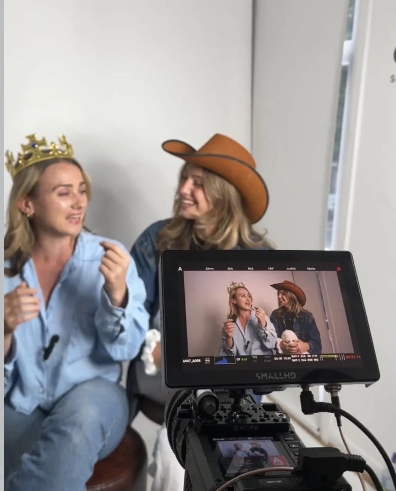 Two women dressed in casual clothing laughing and smiling, one wearing a cowboy hat and pink tinted sunglasses, the other holding a remote, standing in front of a white backdrop.