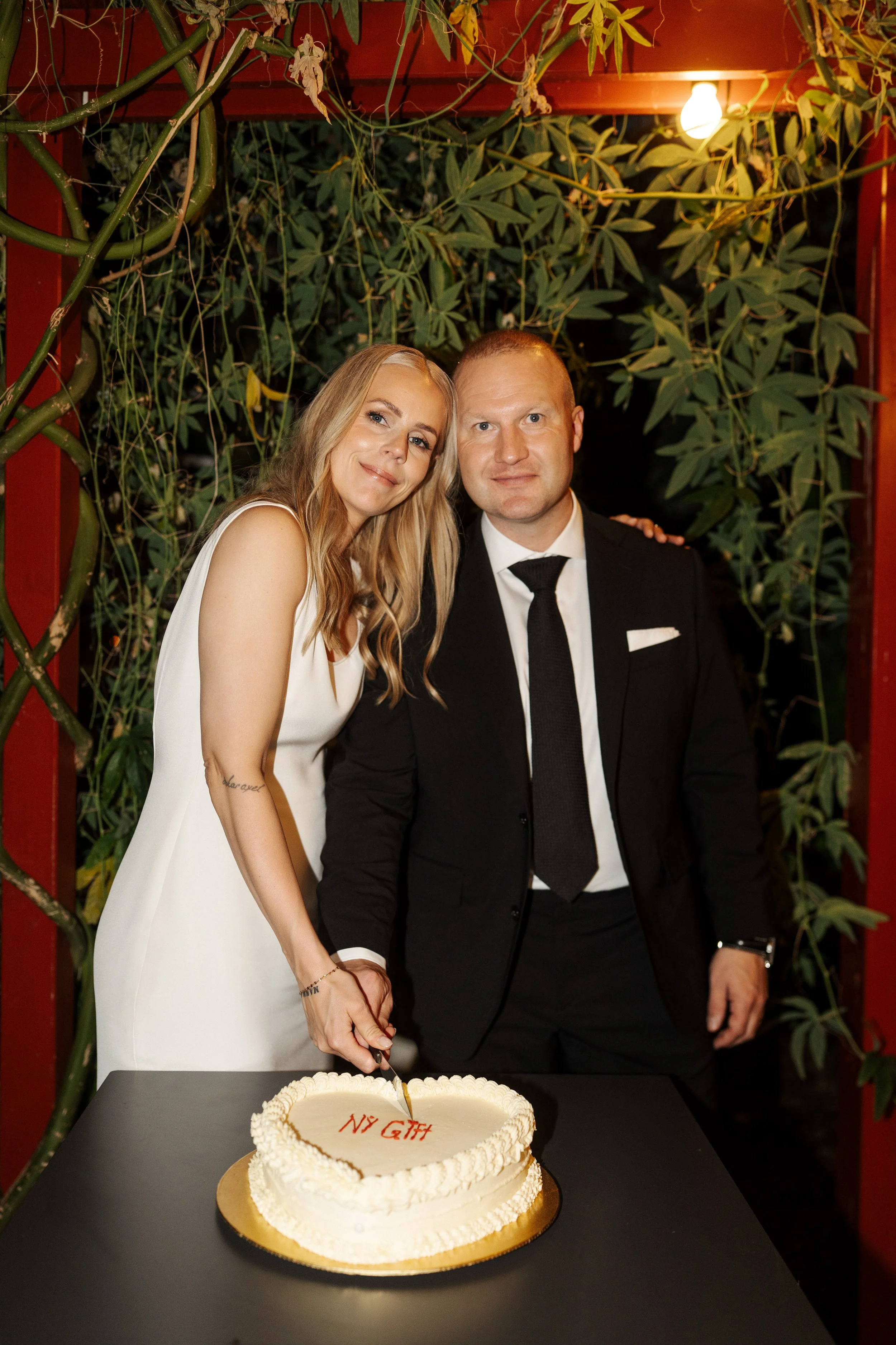 A bride and groom dressed in wedding attire cutting a wedding cake at their celebration. The bride has long blonde hair and a white dress, while the groom is in a black suit with a black tie. They are standing in front of a leafy backdrop with a stri
