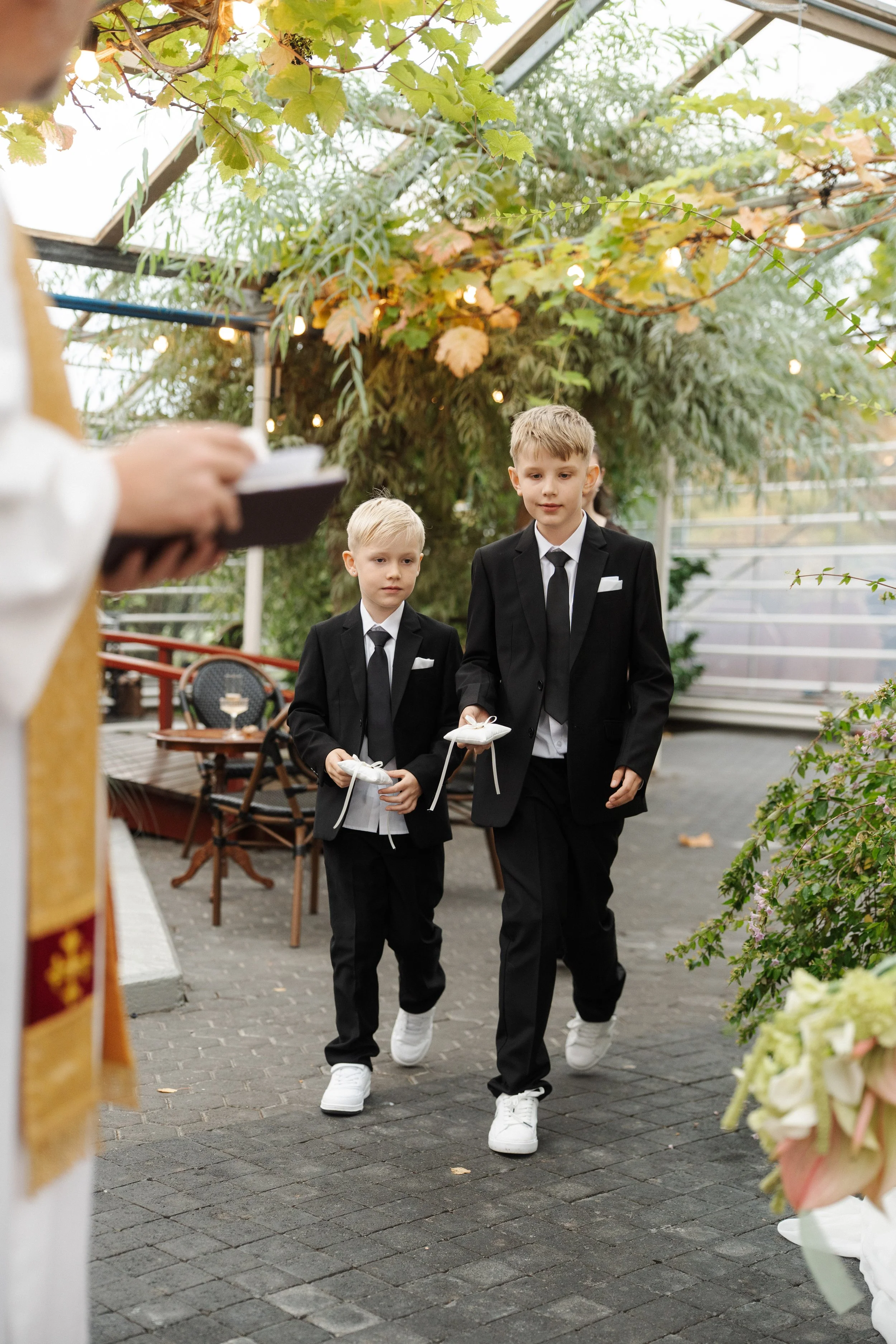 Two boys in black suits and white sneakers participating in a religious ceremony inside a greenhouse, holding small baskets. A person in religious vestments is seen holding a book in the foreground, and there are floral arrangements and outdoor furni