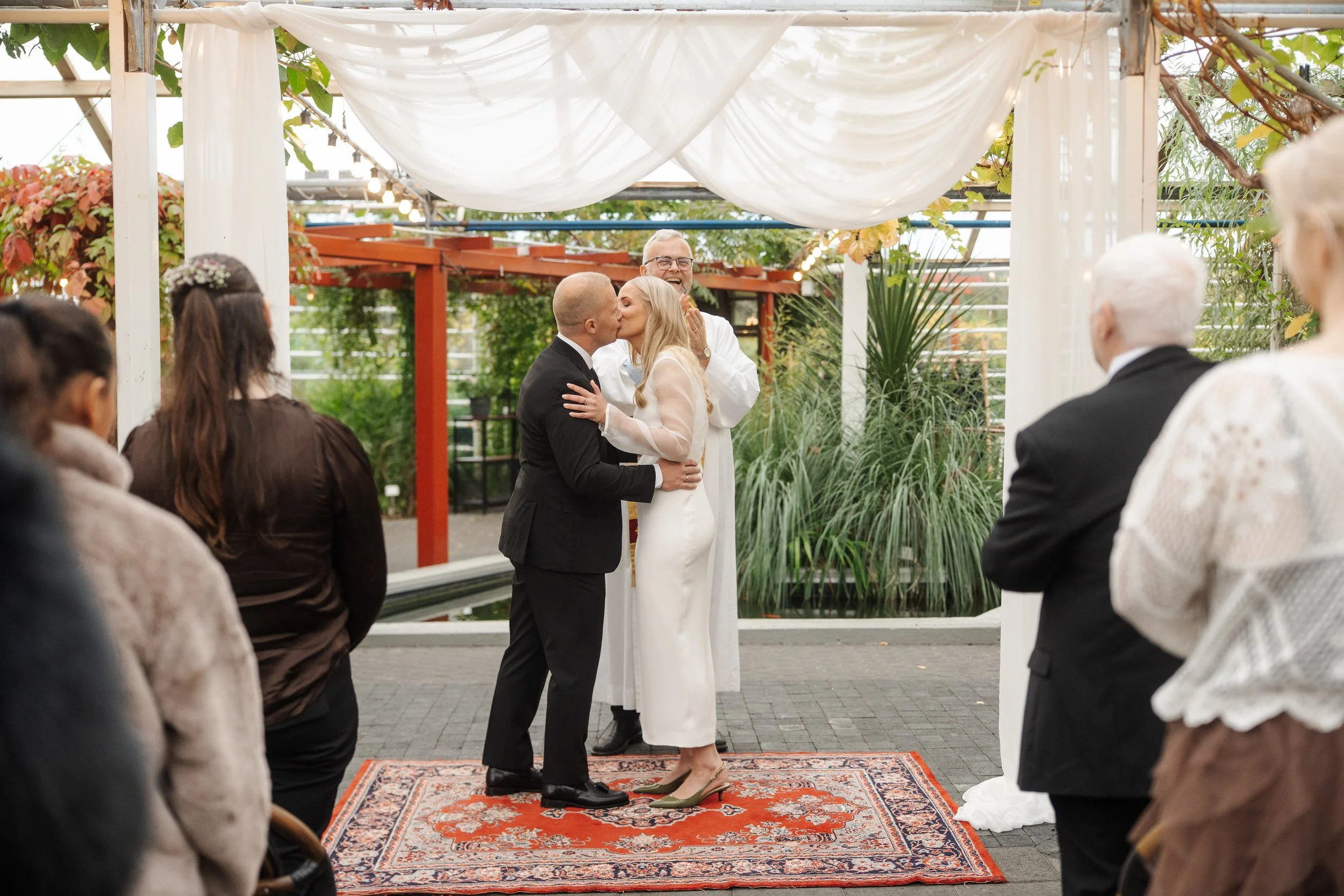 A wedding ceremony with a couple kissing under a white fabric canopy, officiated by a person in a white robe, while guests stand around and watch. The setting is outdoors with plants and string lights in the background.