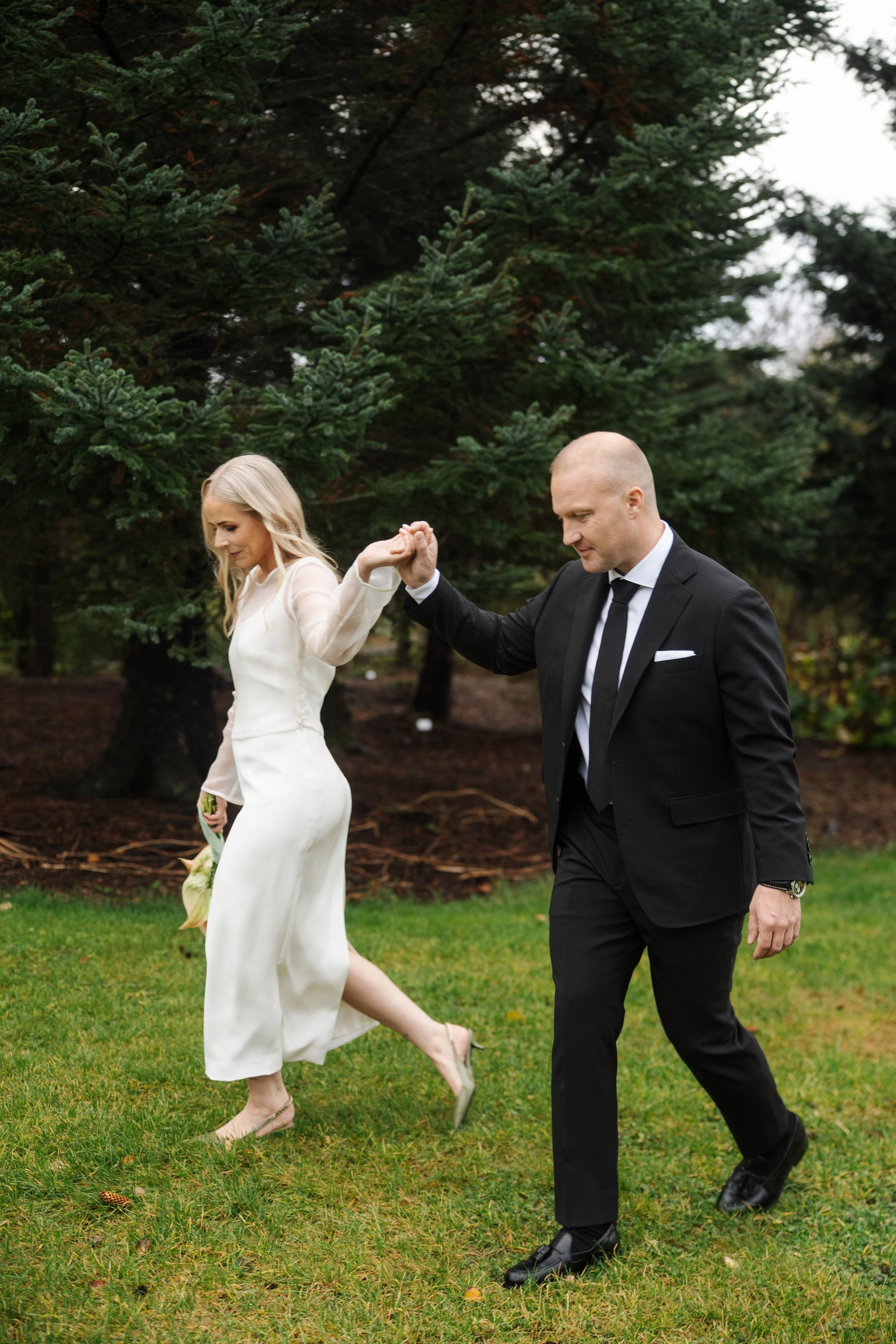 A bride and groom holding hands outdoors, walking on grass with pine trees in the background during daytime.
