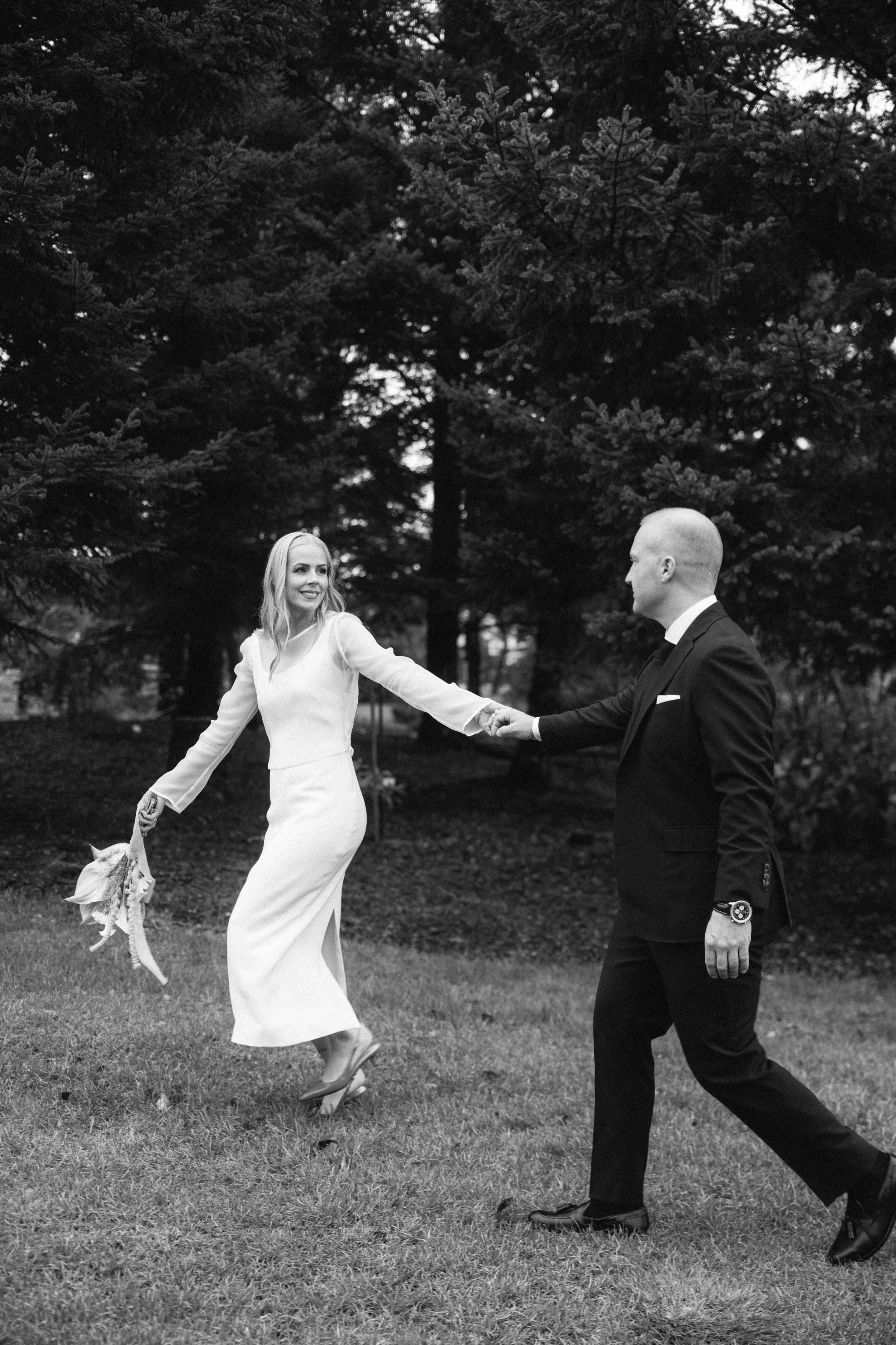 A black-and-white photo of a couple, dressed in wedding attire, holding hands and smiling in a park with large trees in the background.