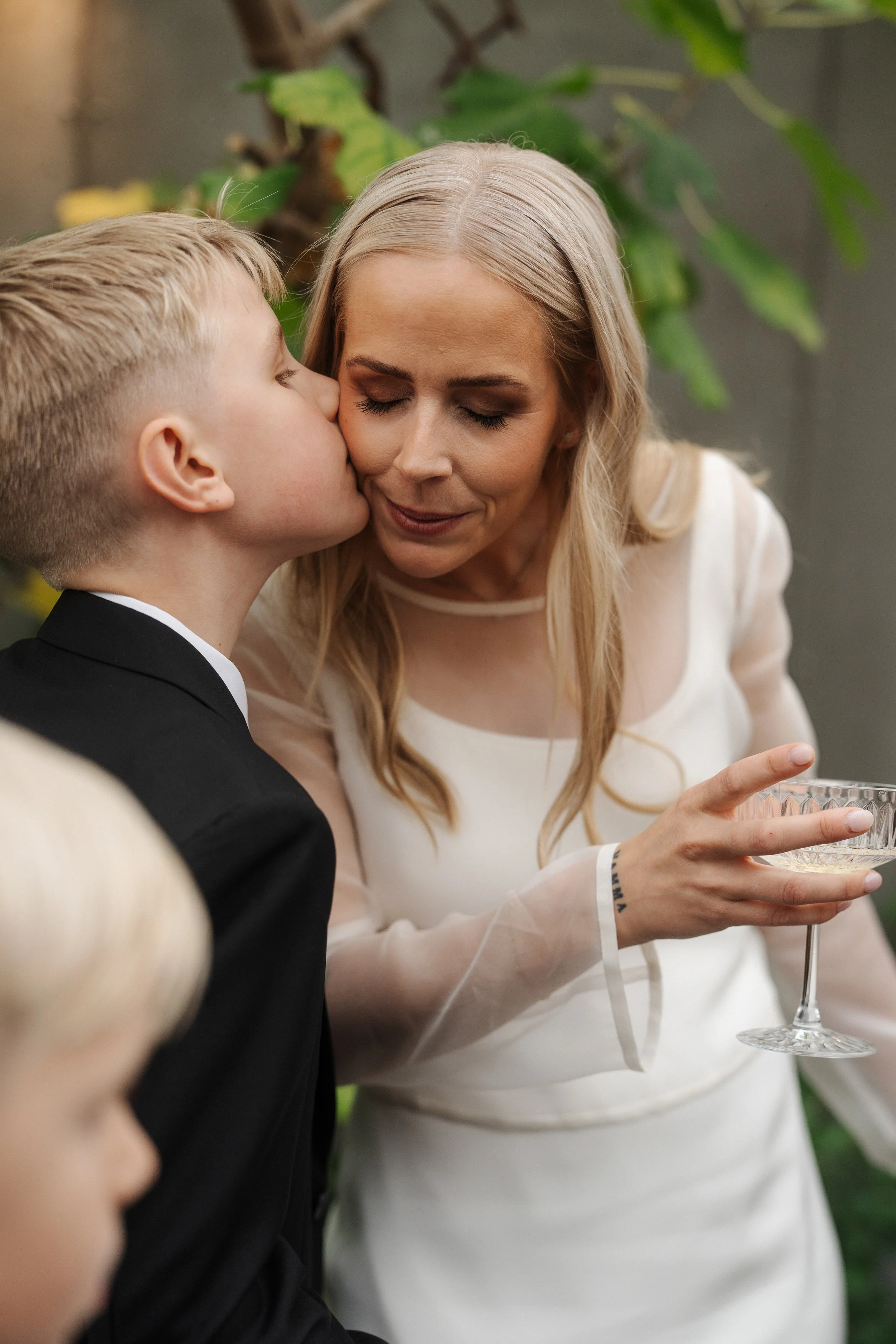 A woman in a white dress is receiving a kiss on the cheek from a young boy dressed in a black suit, during a social gathering or celebration.