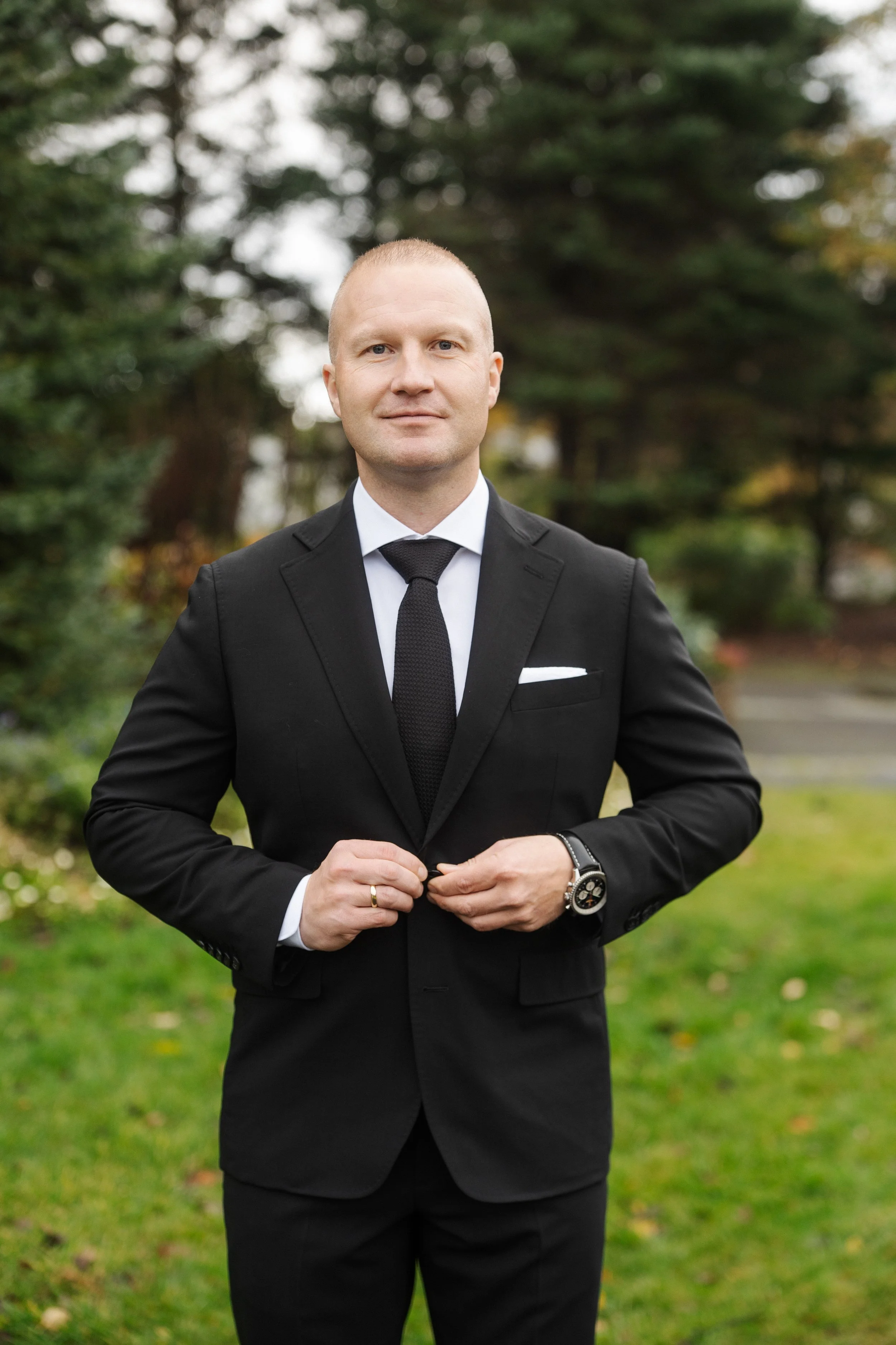A man in a black suit, white shirt, and black tie standing outdoors on a grassy area with trees in the background.