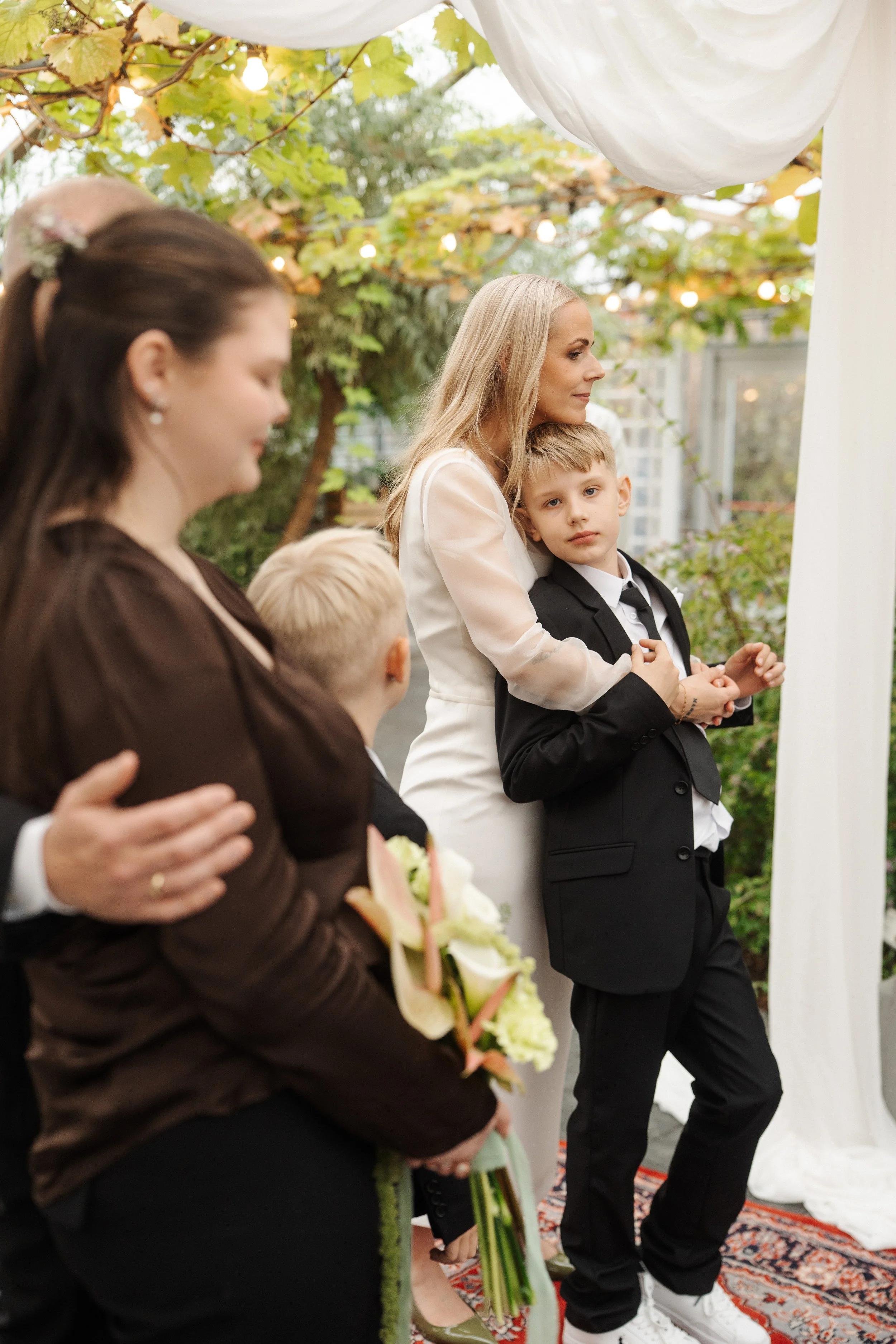 A group of adults and children at an outdoor wedding, with a woman in a white dress holding a young boy leaning on her. There is greenery, string lights, and a decorative white canopy.