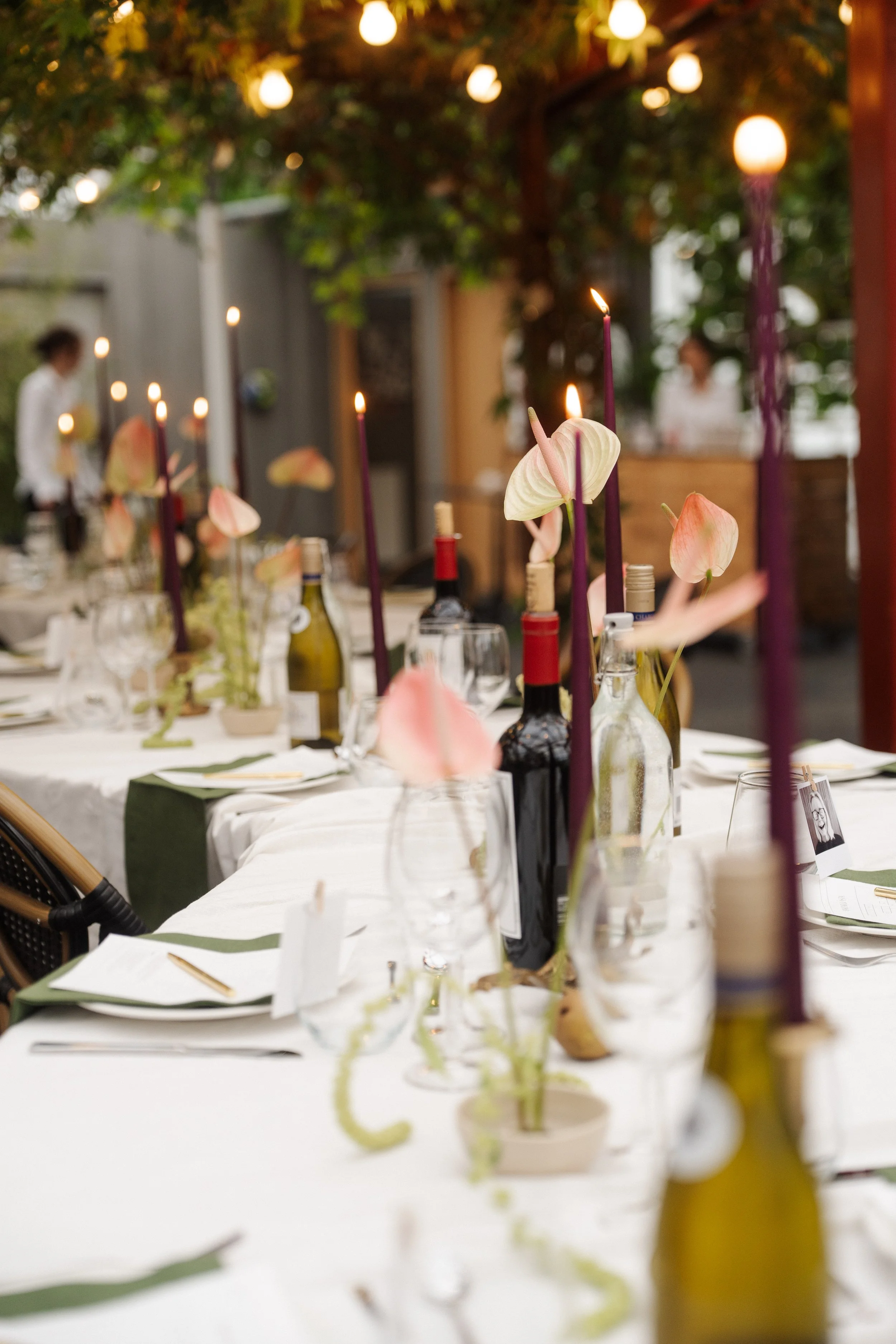 A decorated outdoor dining table with wine bottles, candles, and floral arrangements, set for a special event in a garden or patio area, with trees and string lights overhead.