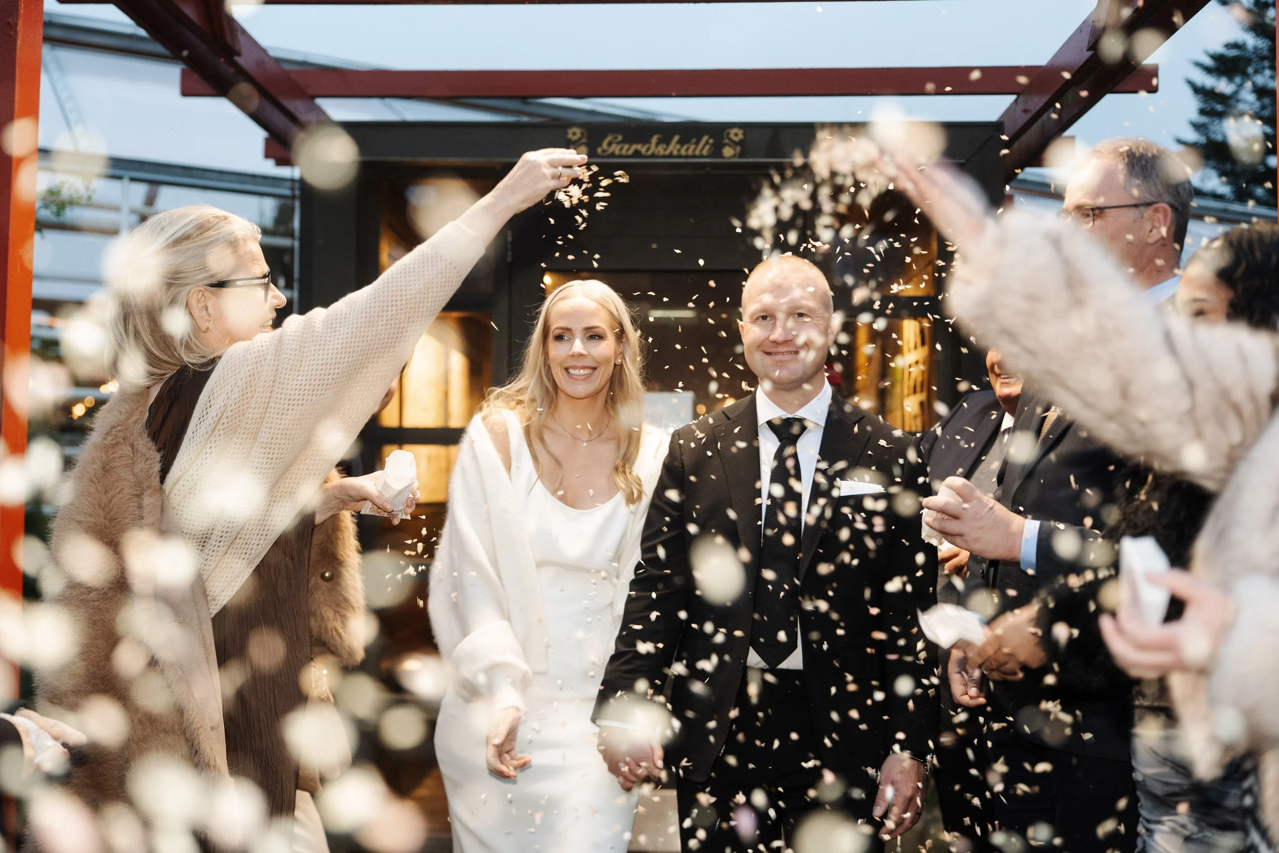 A couple on their wedding day holding hands and smiling, surrounded by friends and family throwing confetti at sunset.