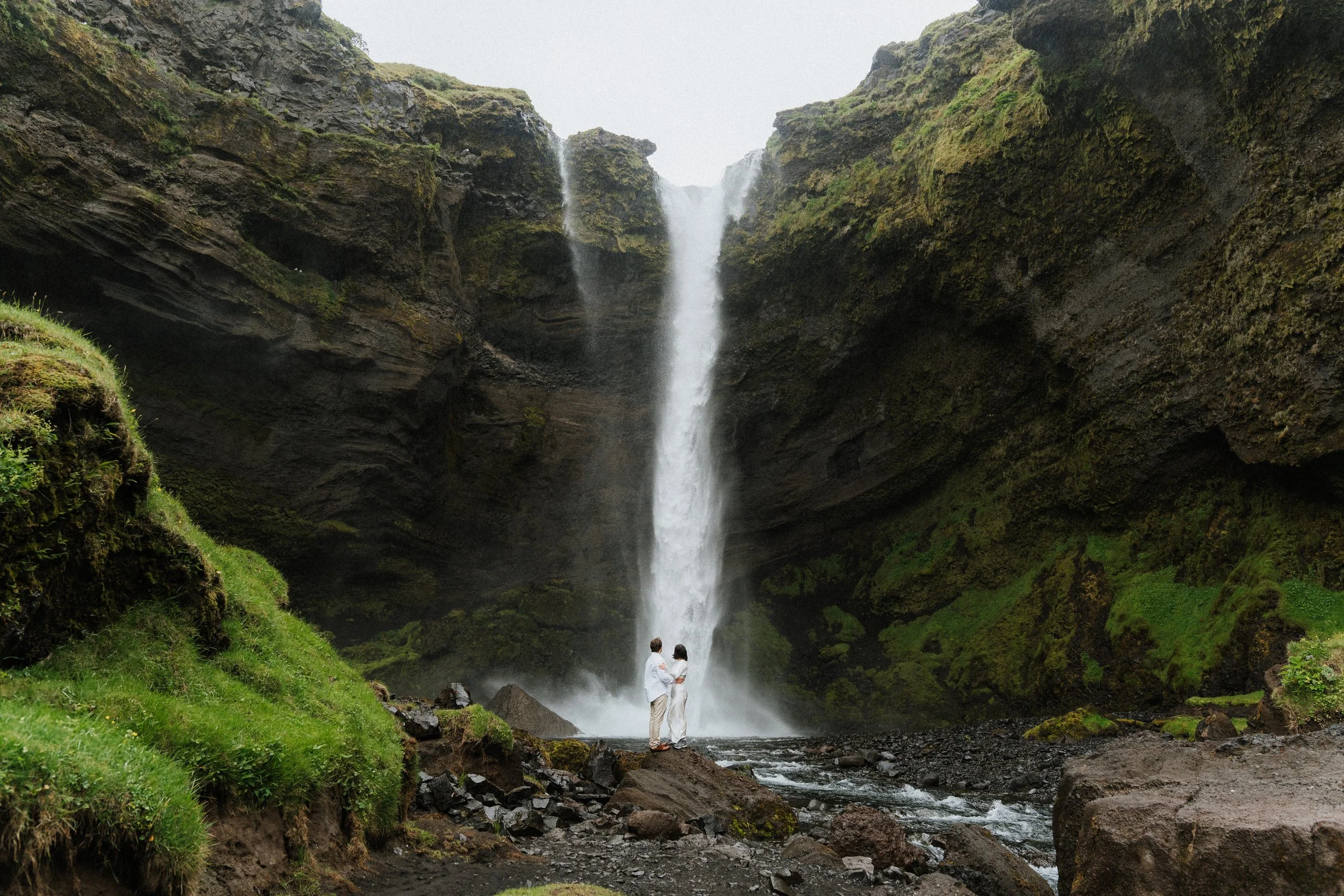 Two people in white outfits stand by a waterfall in a lush, green canyon with moss-covered rocks and flowing water.