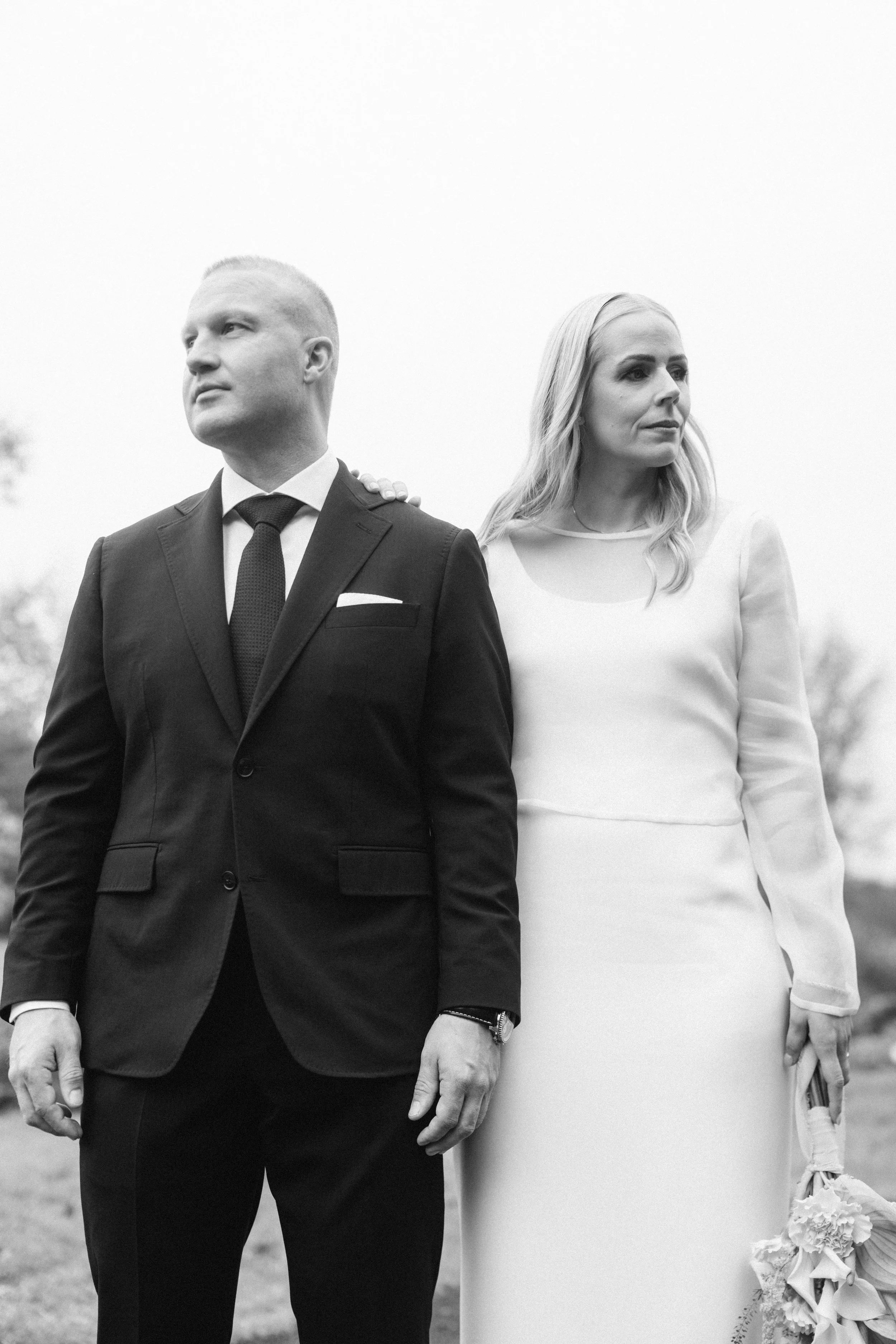 A black and white photo of a man in a dark suit and tie standing next to a woman in a light-colored dress holding a bouquet, outdoors with a blurred background.