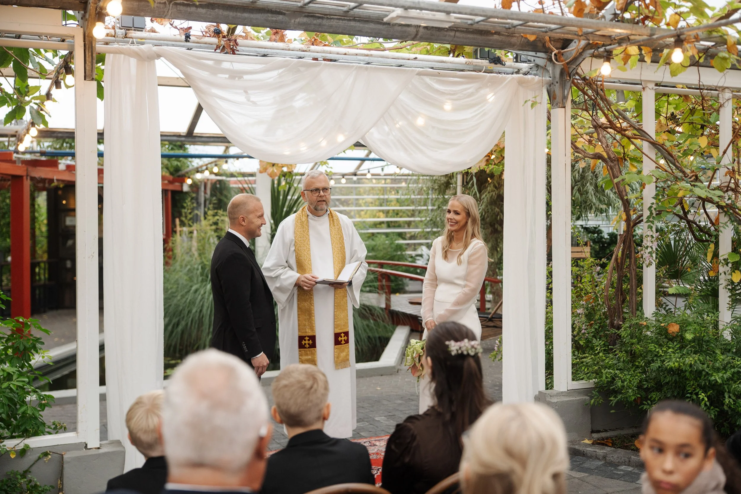 A wedding ceremony outdoors with a priest officiating, a bride in white holding a bouquet, and a groom in a black suit. Guests are seated, and the setting is decorated with white drapes and string lights.