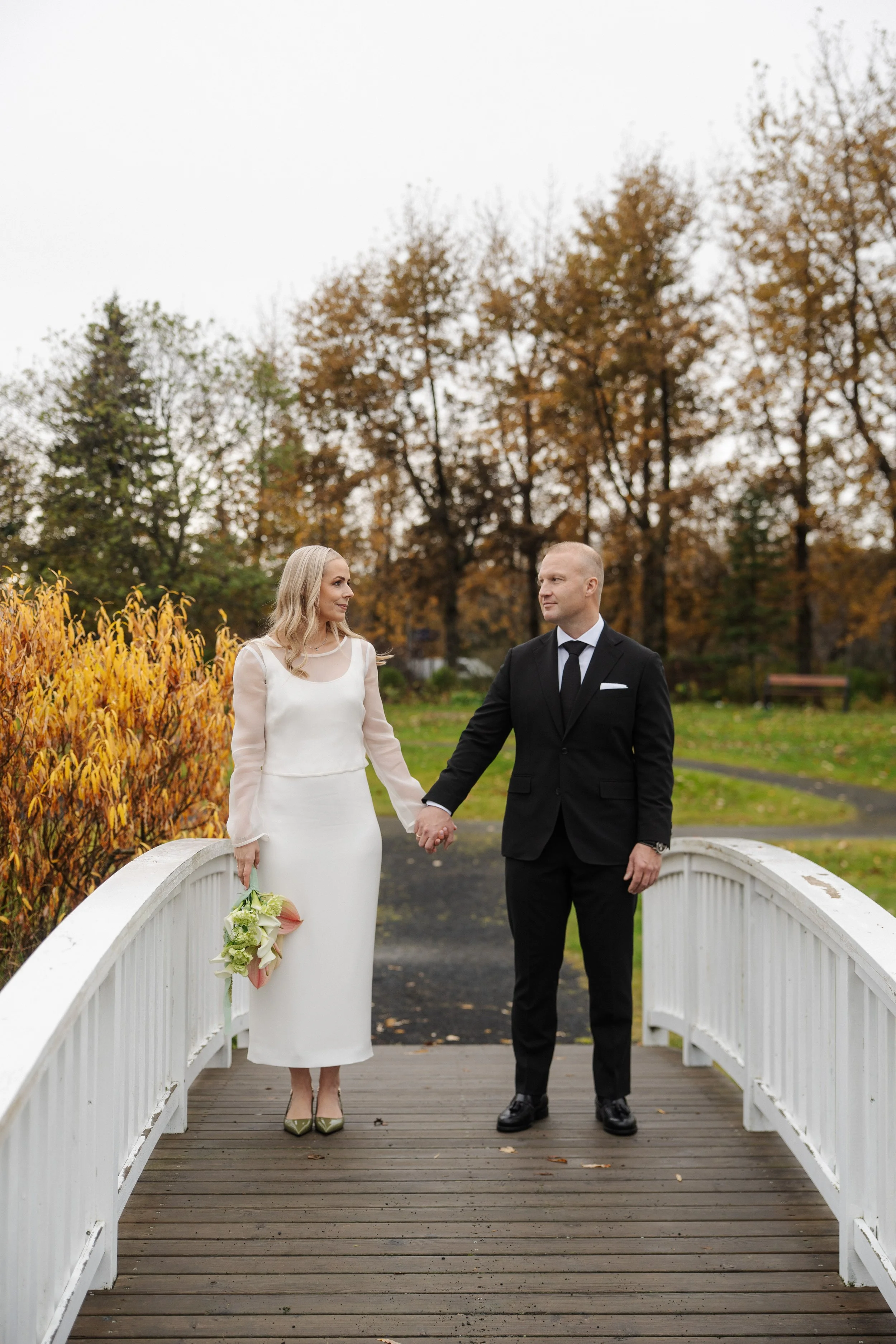 A bride and groom holding hands on a small white bridge in an outdoor park with fall foliage.