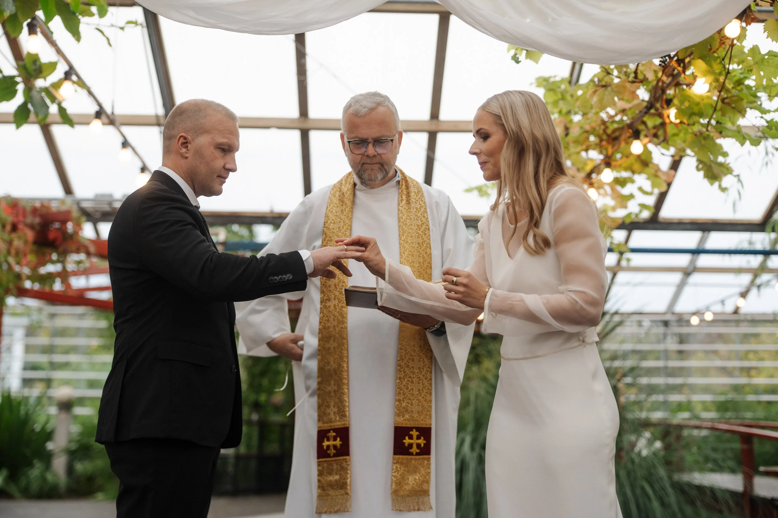A wedding ceremony with a groom placing a ring on the bride's finger, officiated by a priest in a bright greenhouse setting surrounded by plants and string lights.