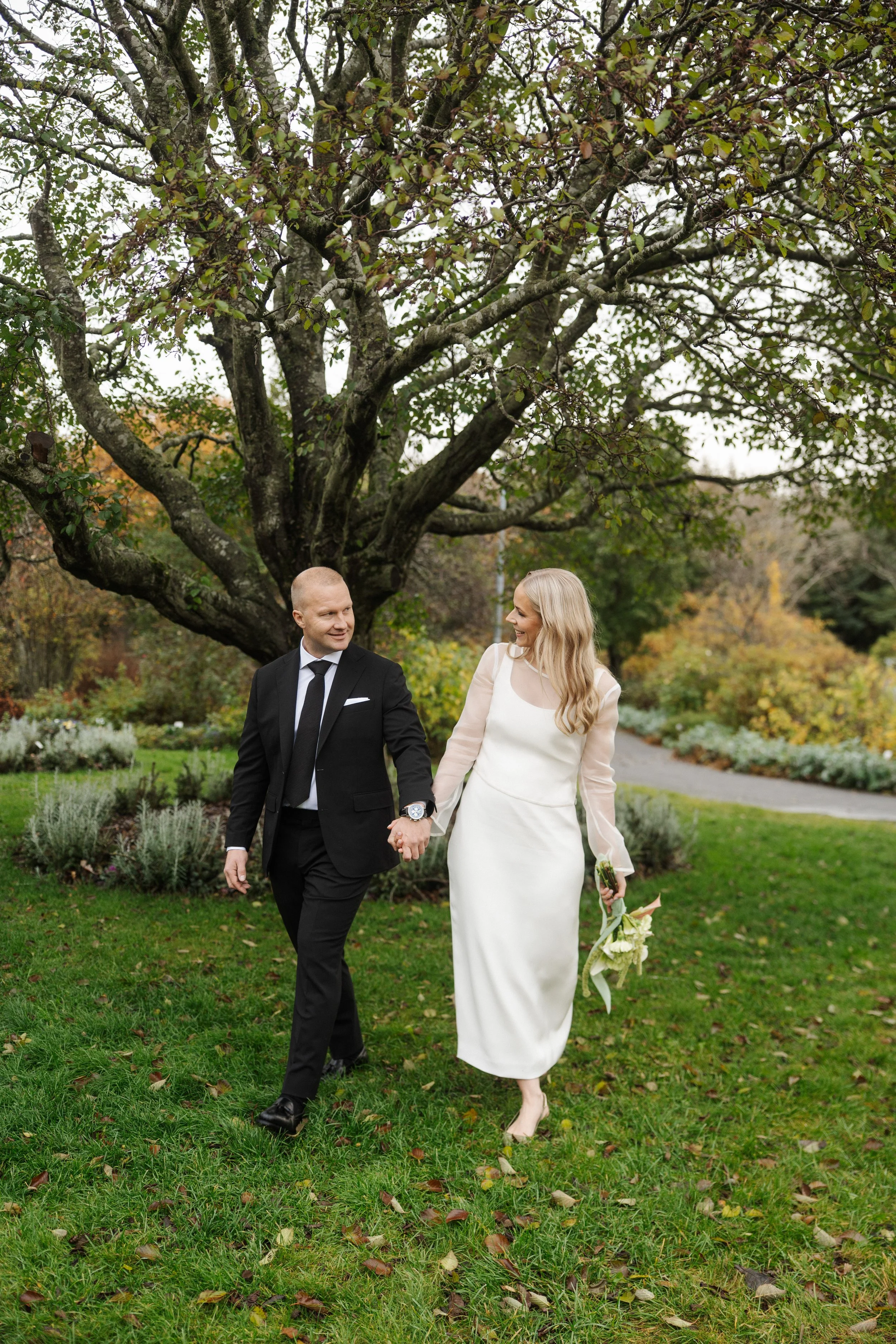 A blonde woman in a long white dress holding a bouquet of flowers and a man in a black suit and tie walking hand in hand under a large tree during autumn.