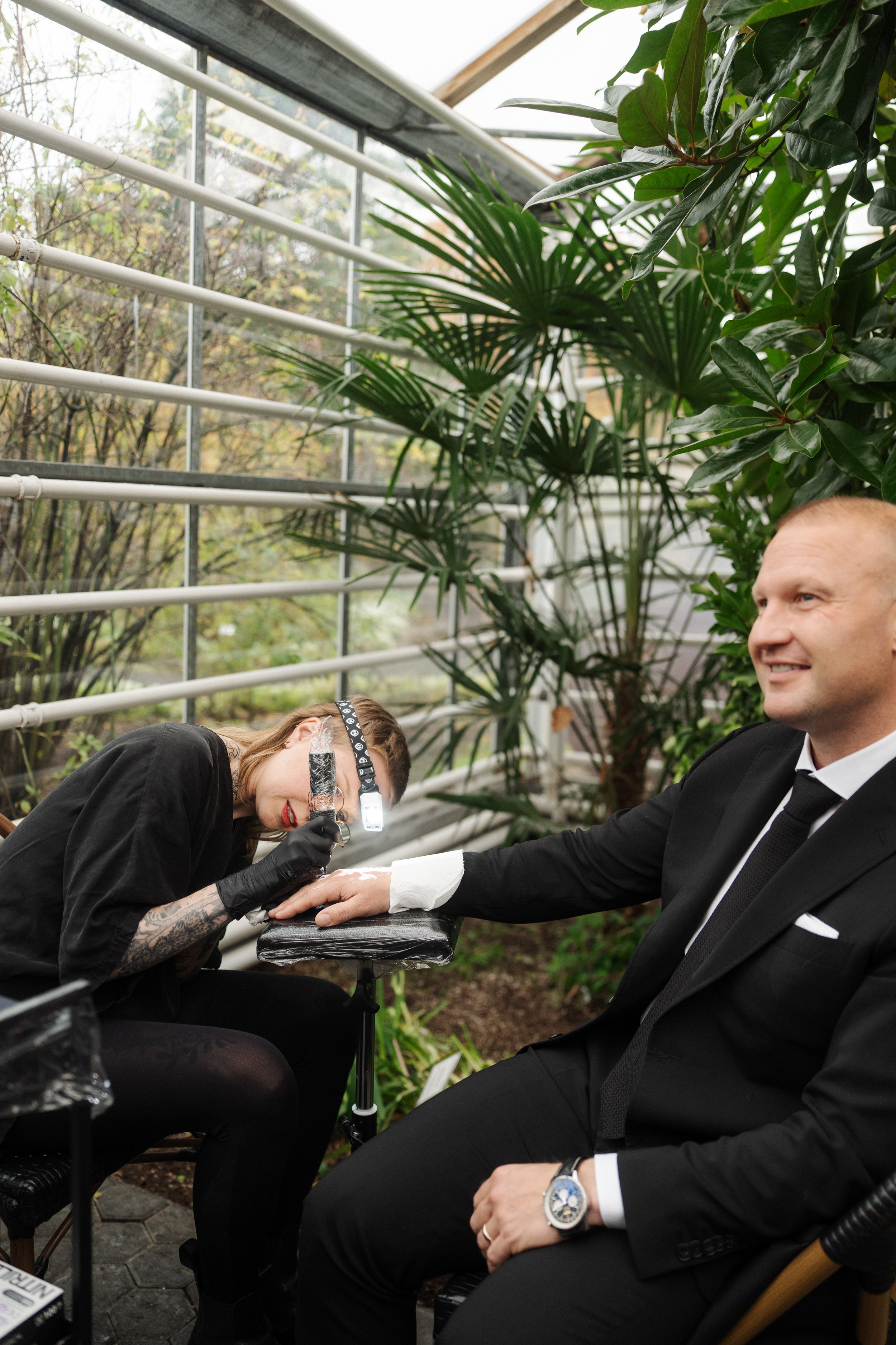 A tattooed woman with glasses and a headlamp gives a manicure to a man in a black suit, seated outdoors among large green plants.