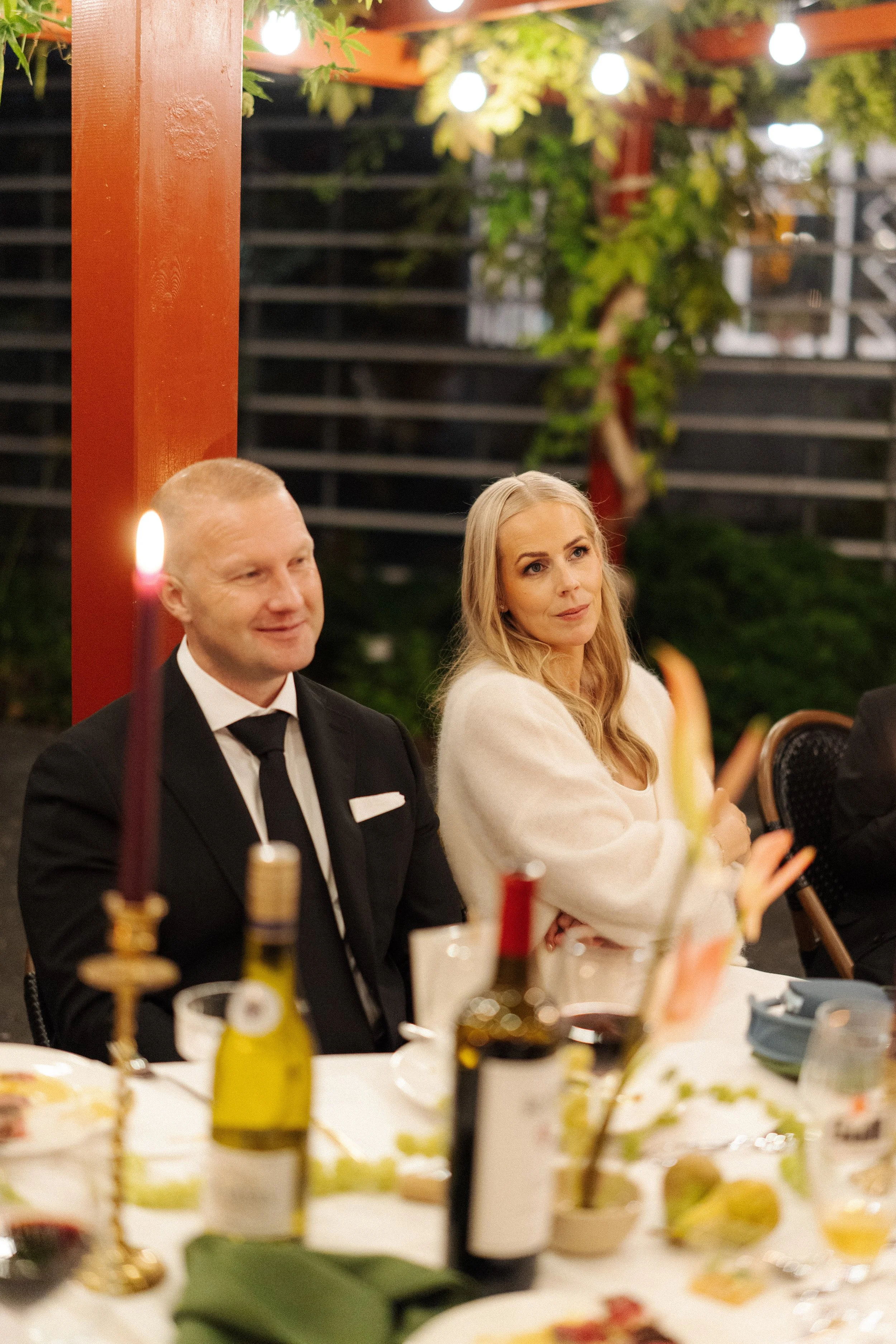 A man and a woman sitting at a dinner table with food, wine, and a lit candle, surrounded by string lights and greenery at night.