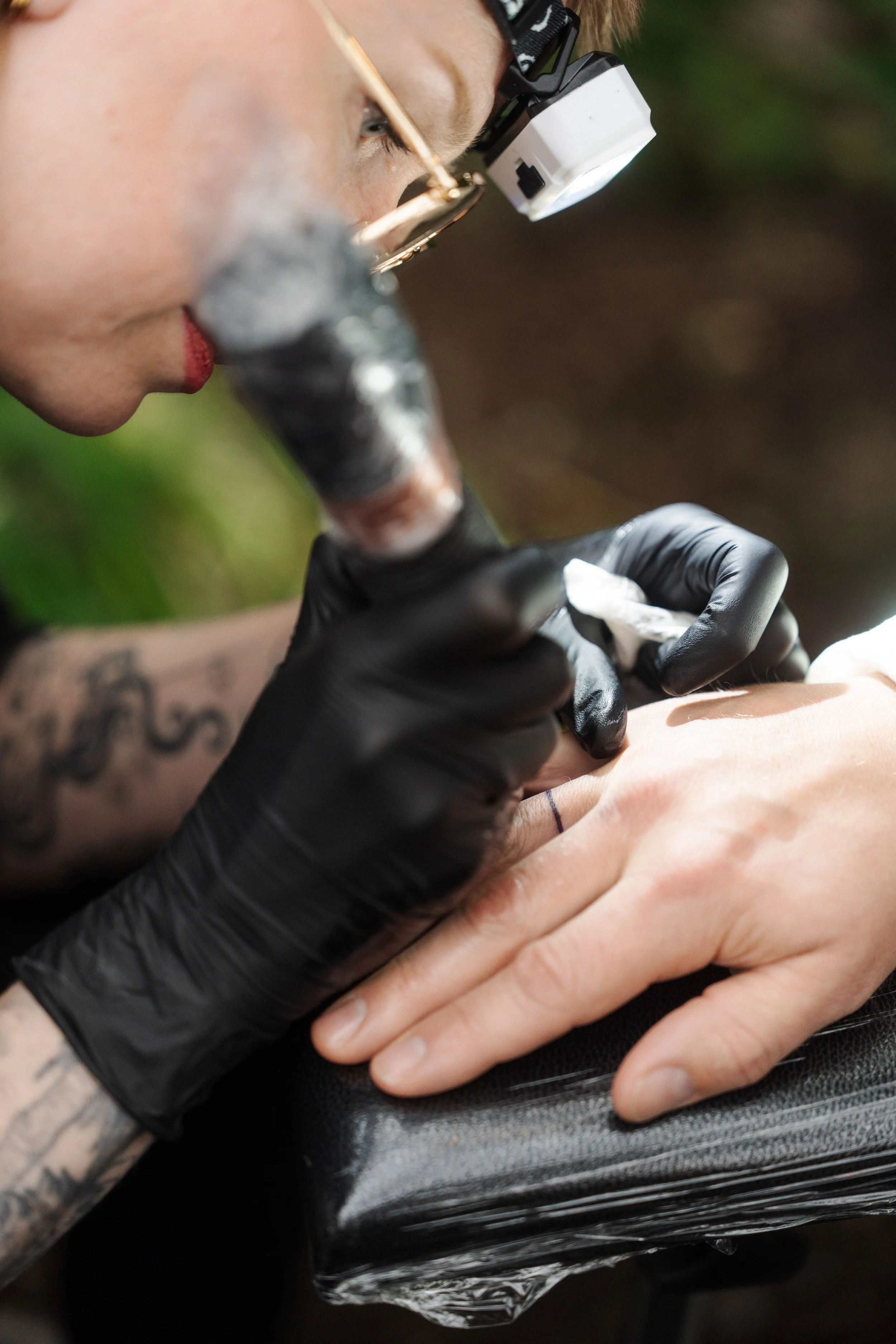 A tattoo artist with black gloves, glasses, and a headlamp tattooing a person's hand with a black ink tattoo.
