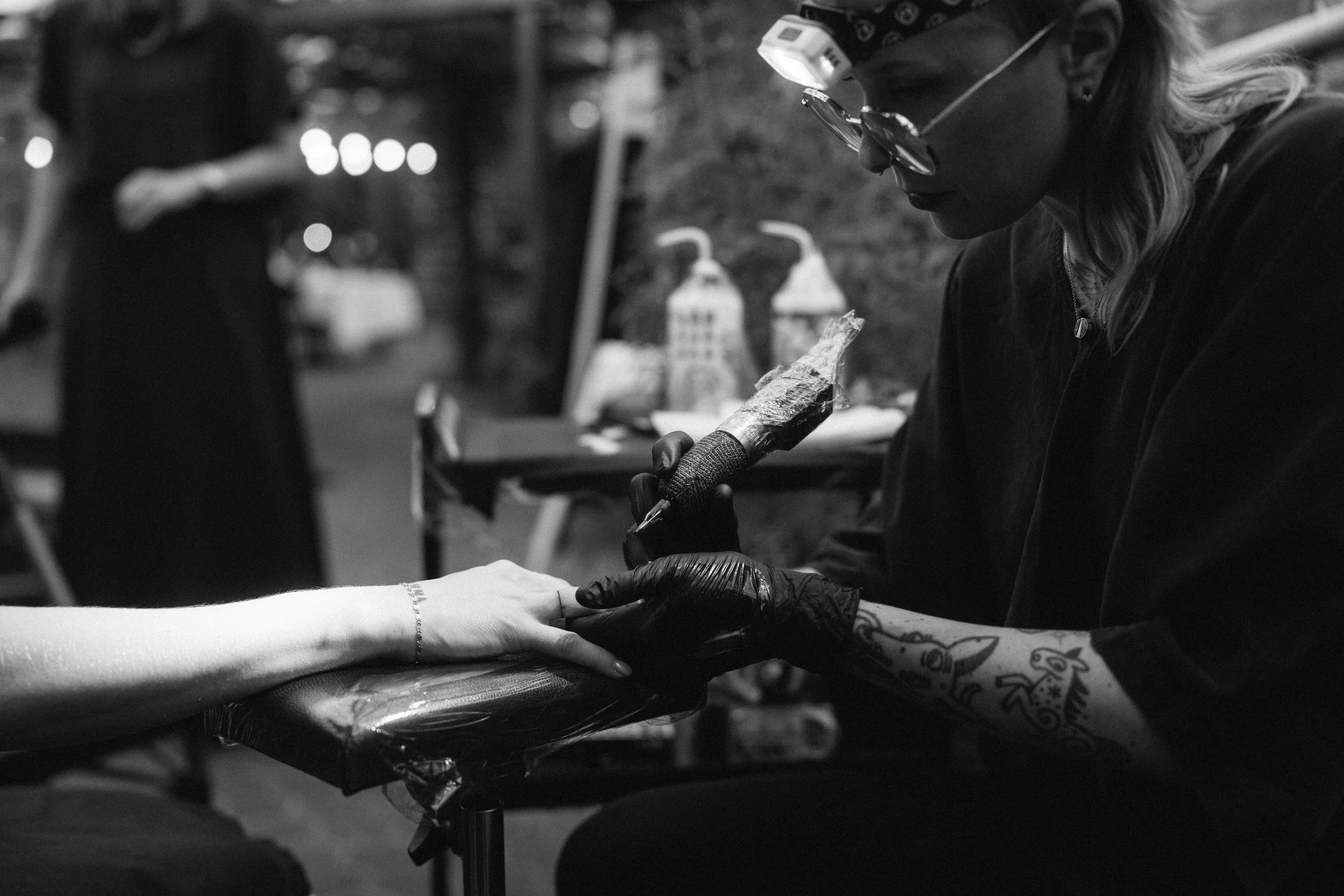 A woman with tattoos on her arm and wearing gloves is giving a manicure to a person with a bracelet on their wrist, in a black and white photo.