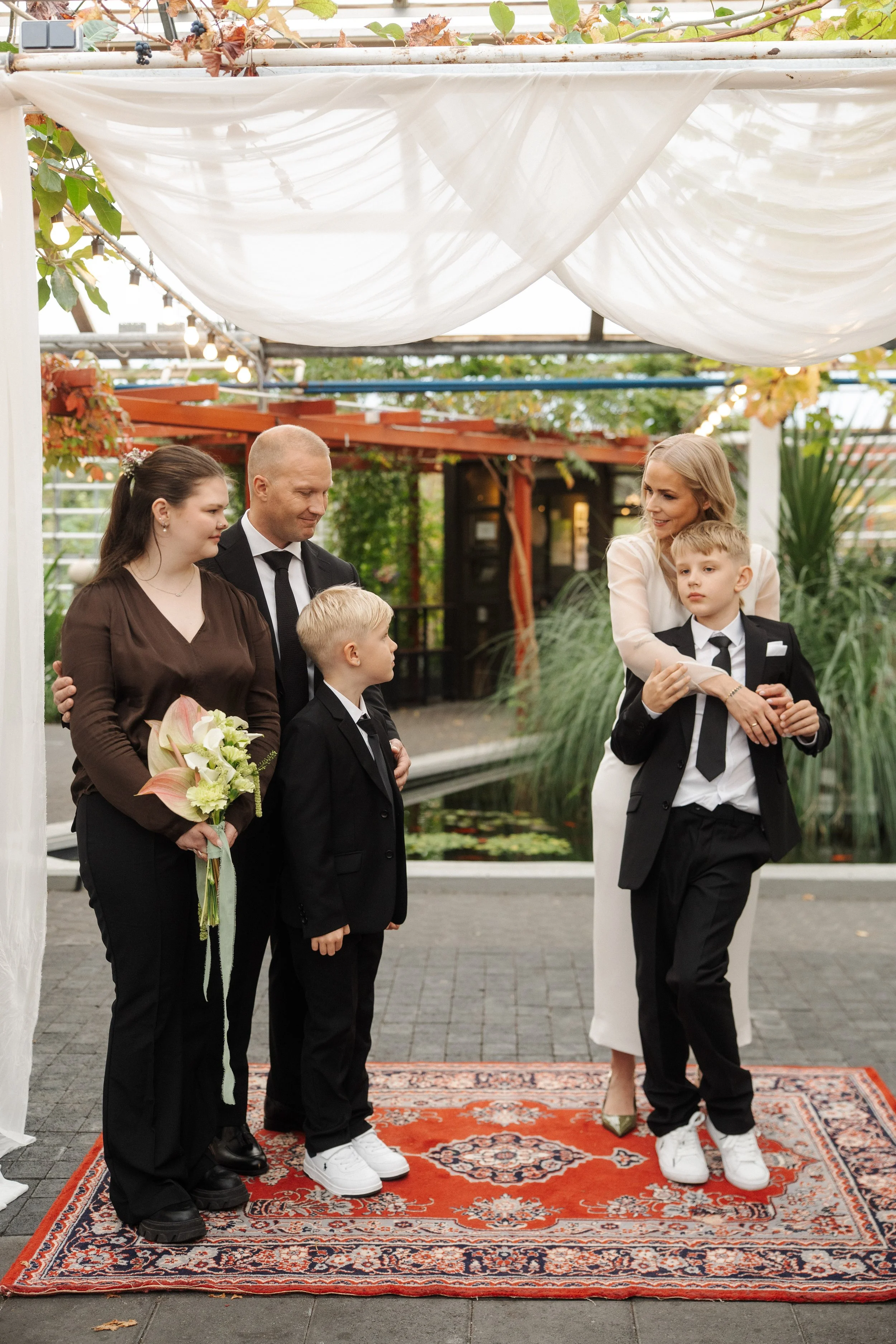 A family gathered for a wedding ceremony outdoors, with a woman in a white dress embracing a young boy in a black suit, while other family members look on.