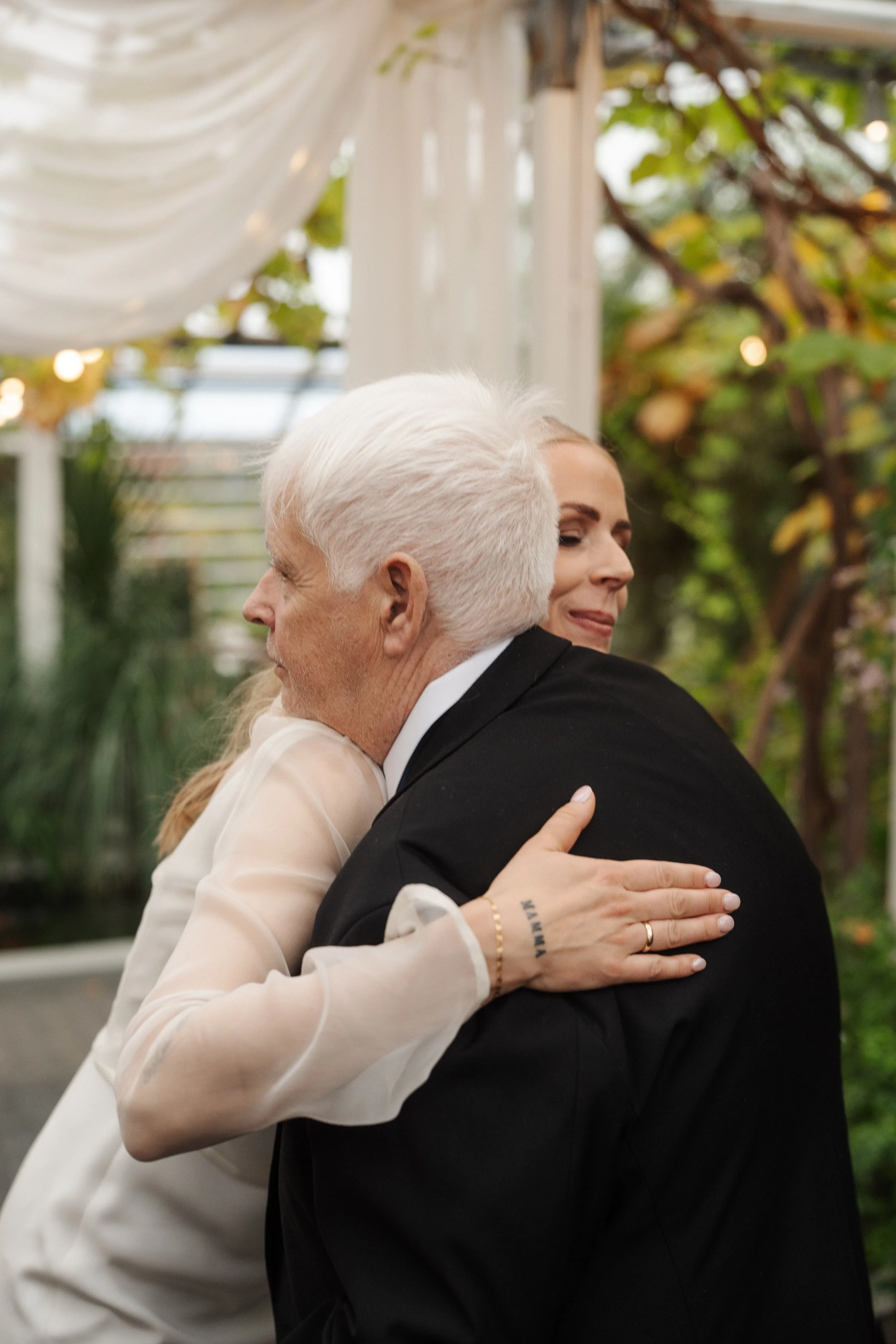 A woman with blonde hair hugging an older man with white hair at an outdoor event, possibly a wedding, with greenery and drapery in the background.