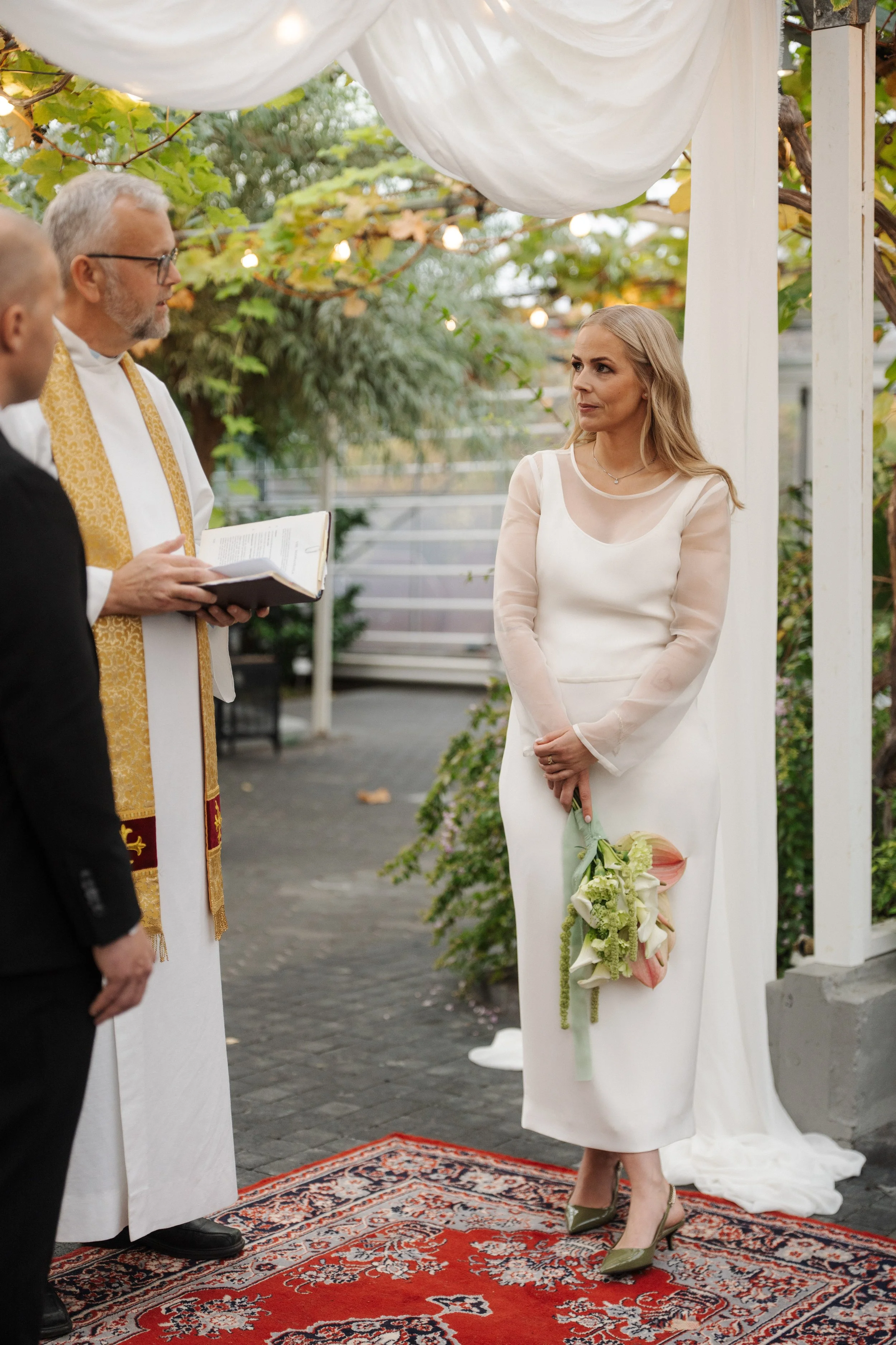 A woman in a white dress holding a bouquet of flowers during a wedding ceremony outdoors, with a priest or officiant and another person partially visible.