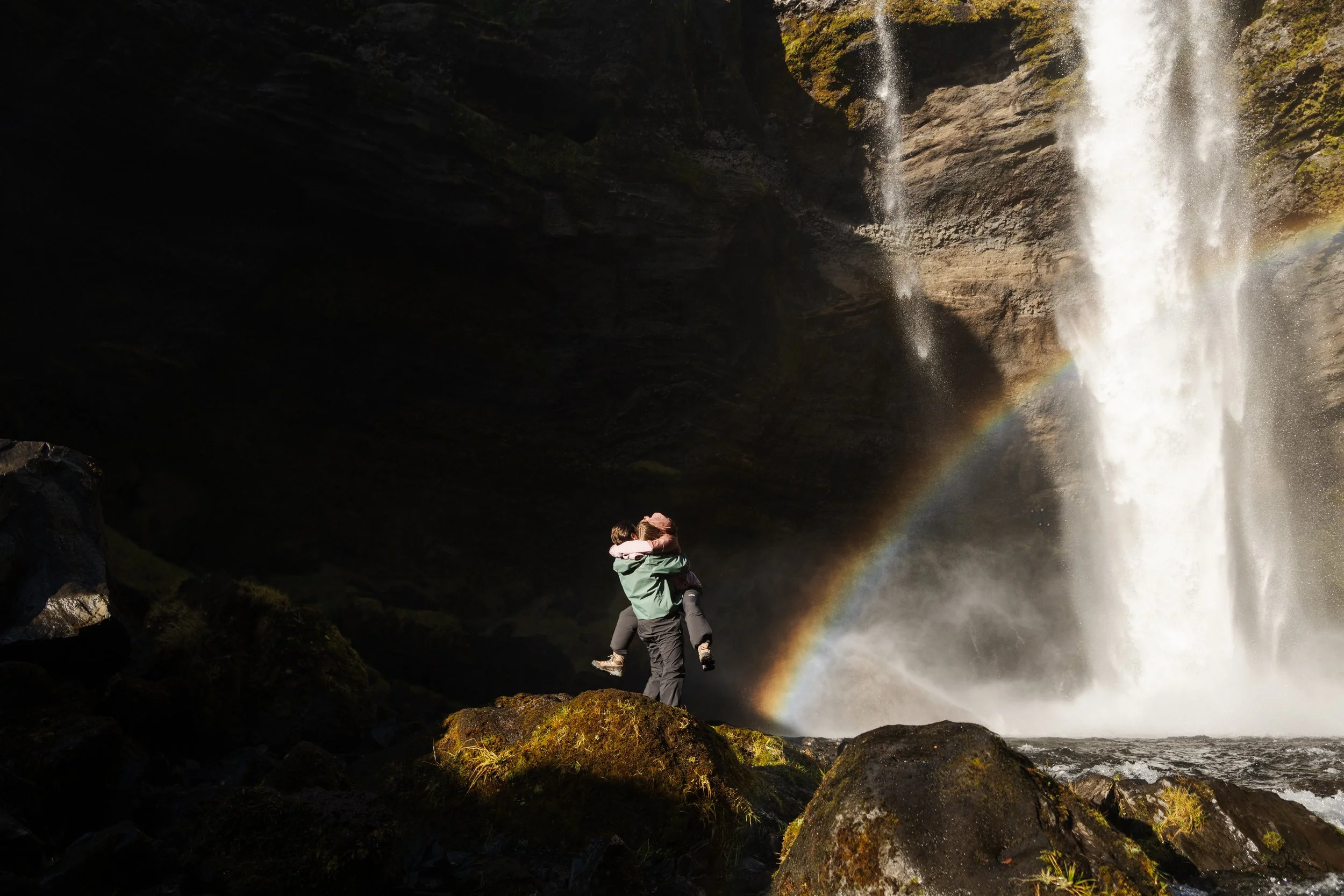 Two people embracing near a waterfall with a rainbow forming in the mist, surrounded by moss-covered rocks and dark cave-like overhang.