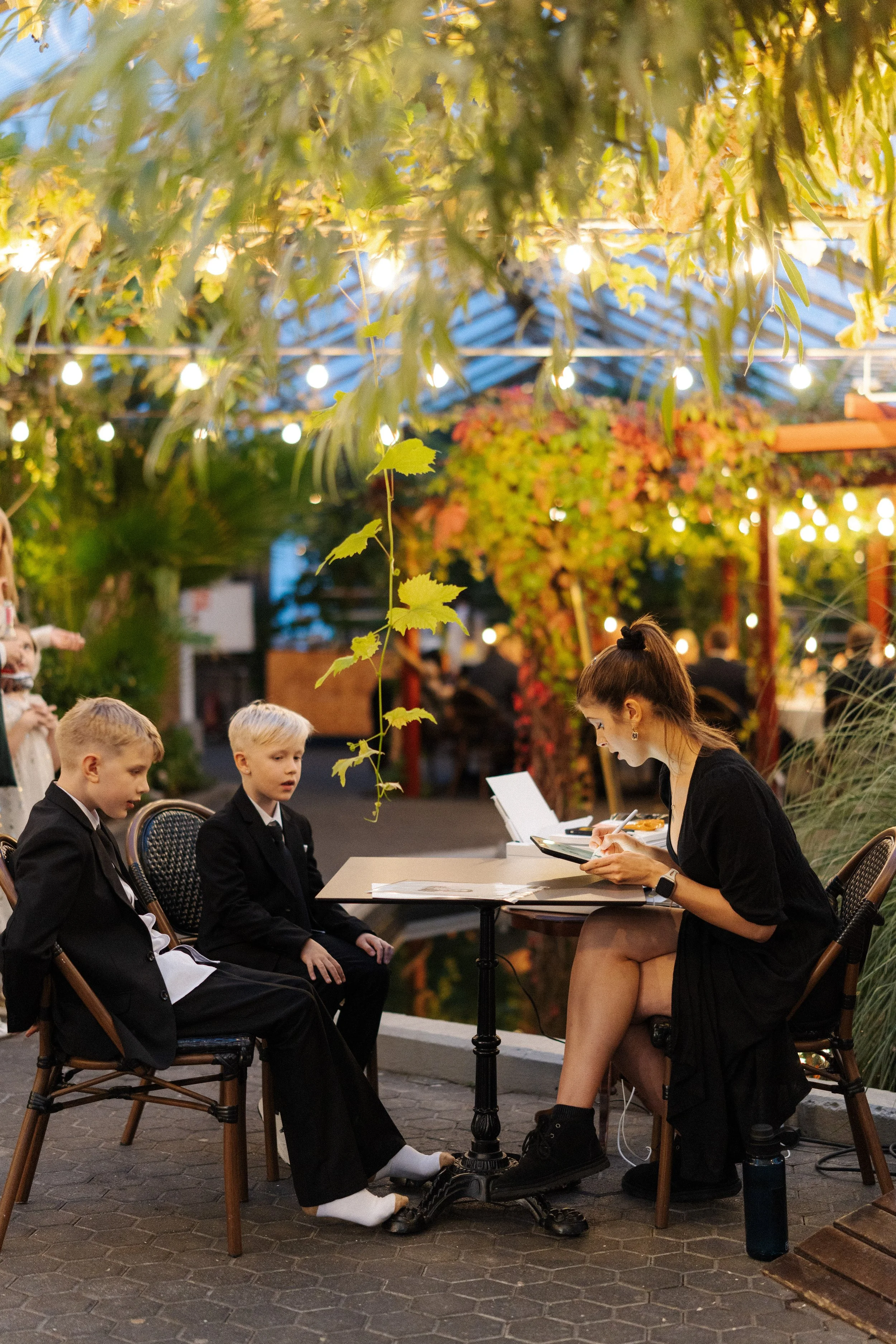 A woman is sitting at a table outdoors, looking at her phone or a menu, with two young boys in suits sitting across from her. The setting is decorated with hanging lights and lush green and red foliage, suggesting an evening in a cozy outdoor dining 