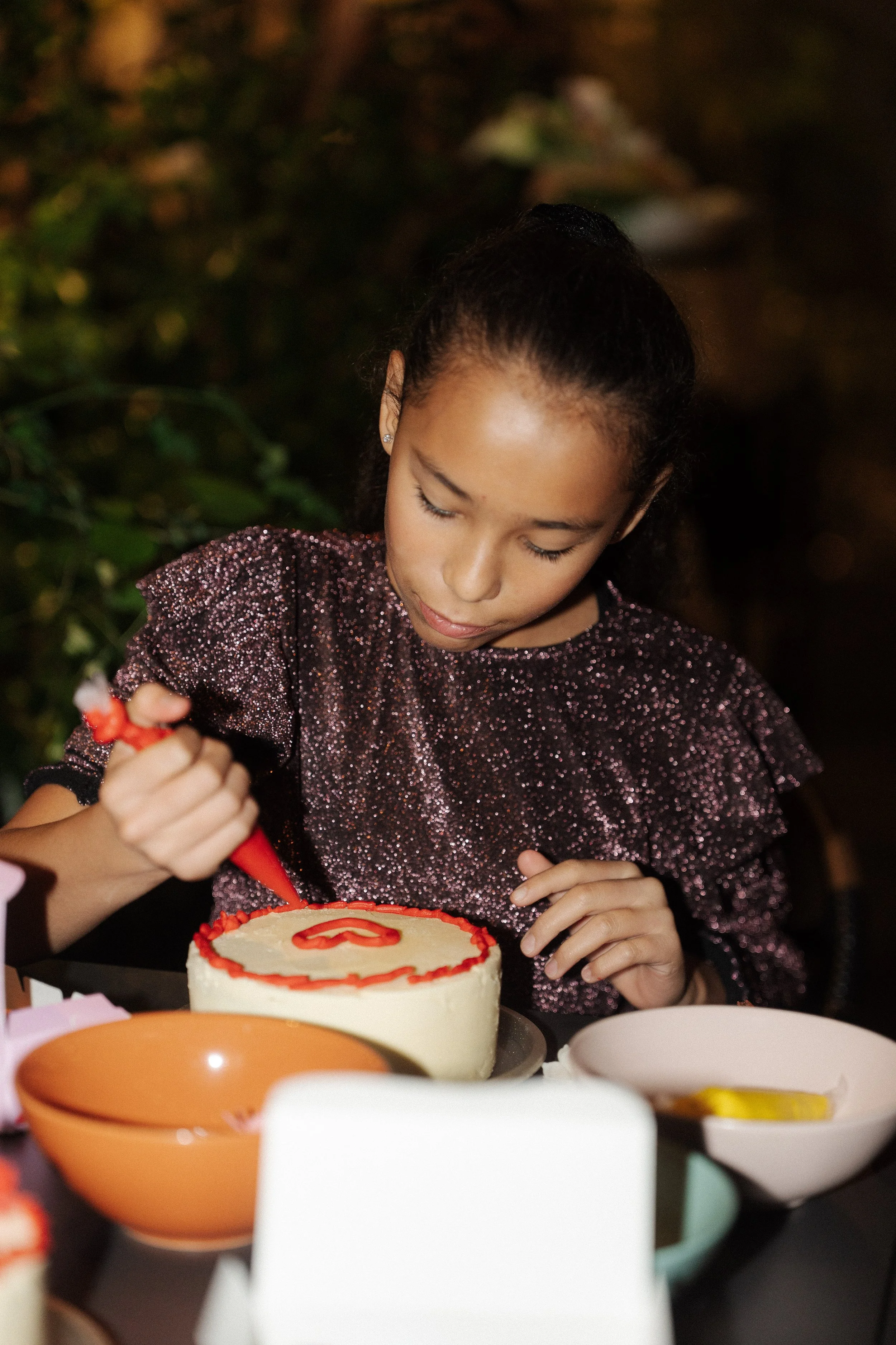 A young girl in a sparkly purple top decorates a white birthday cake with red icing.