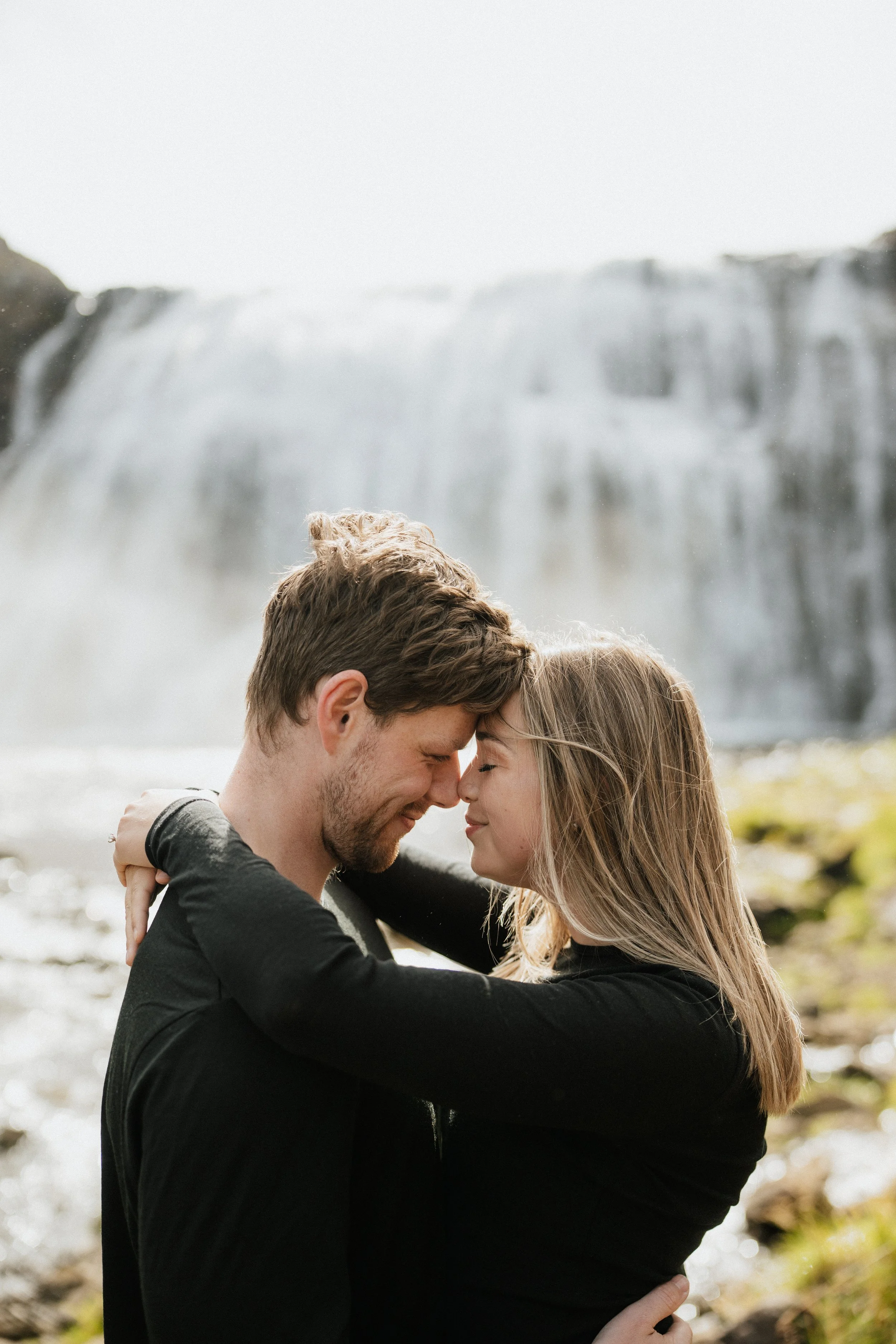 A couple embracing with their foreheads touching outdoors with a waterfall in the background.