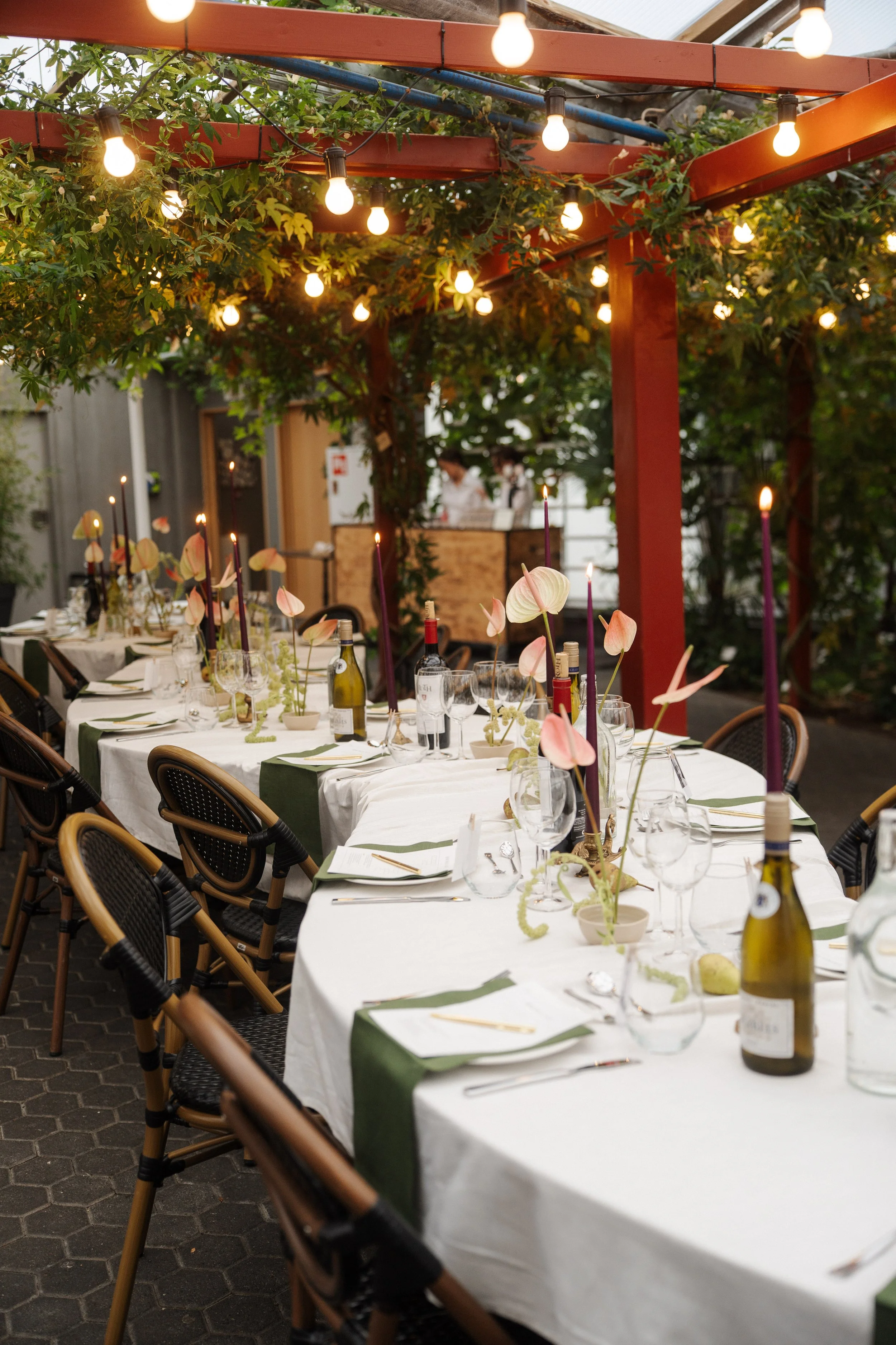 A long outdoor dining table decorated with wine bottles, glasses, flowers, and candles, set under string lights and surrounded by greenery.