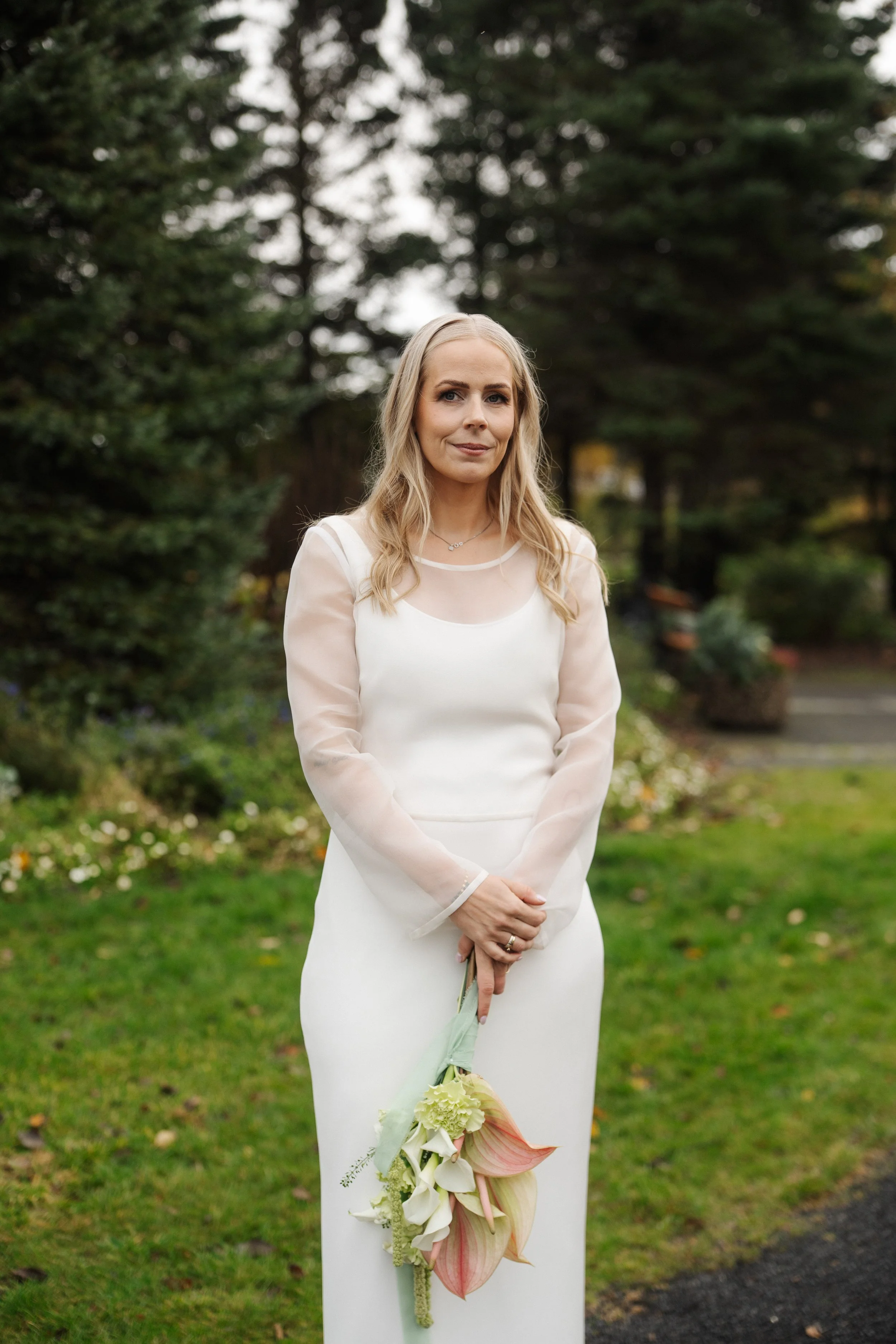 A woman in a white dress holding a bouquet of flowers standing outdoors in a garden with trees and plants.