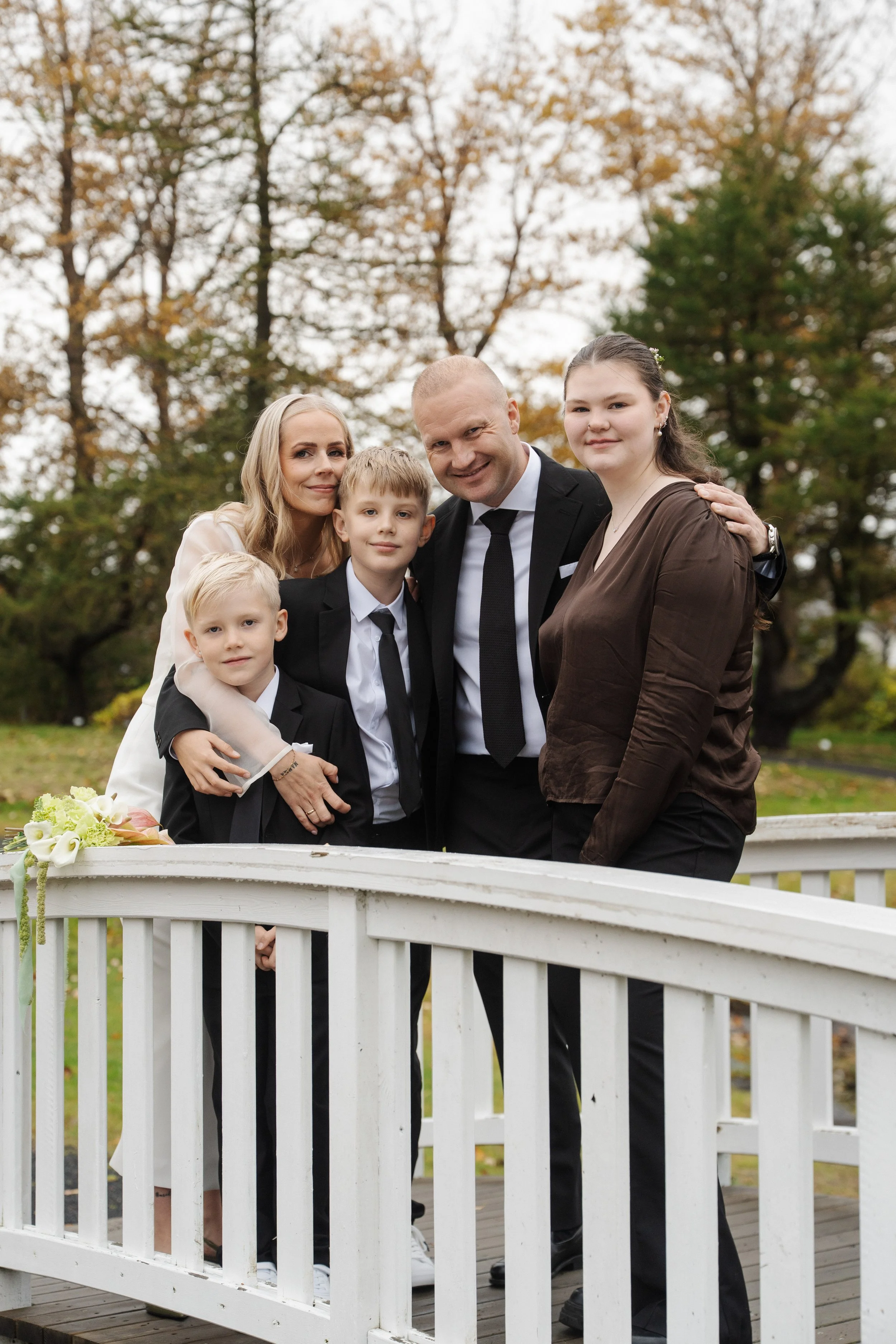 Family gathered outdoors on a small white bridge, posing for a photo in autumn with trees in the background.