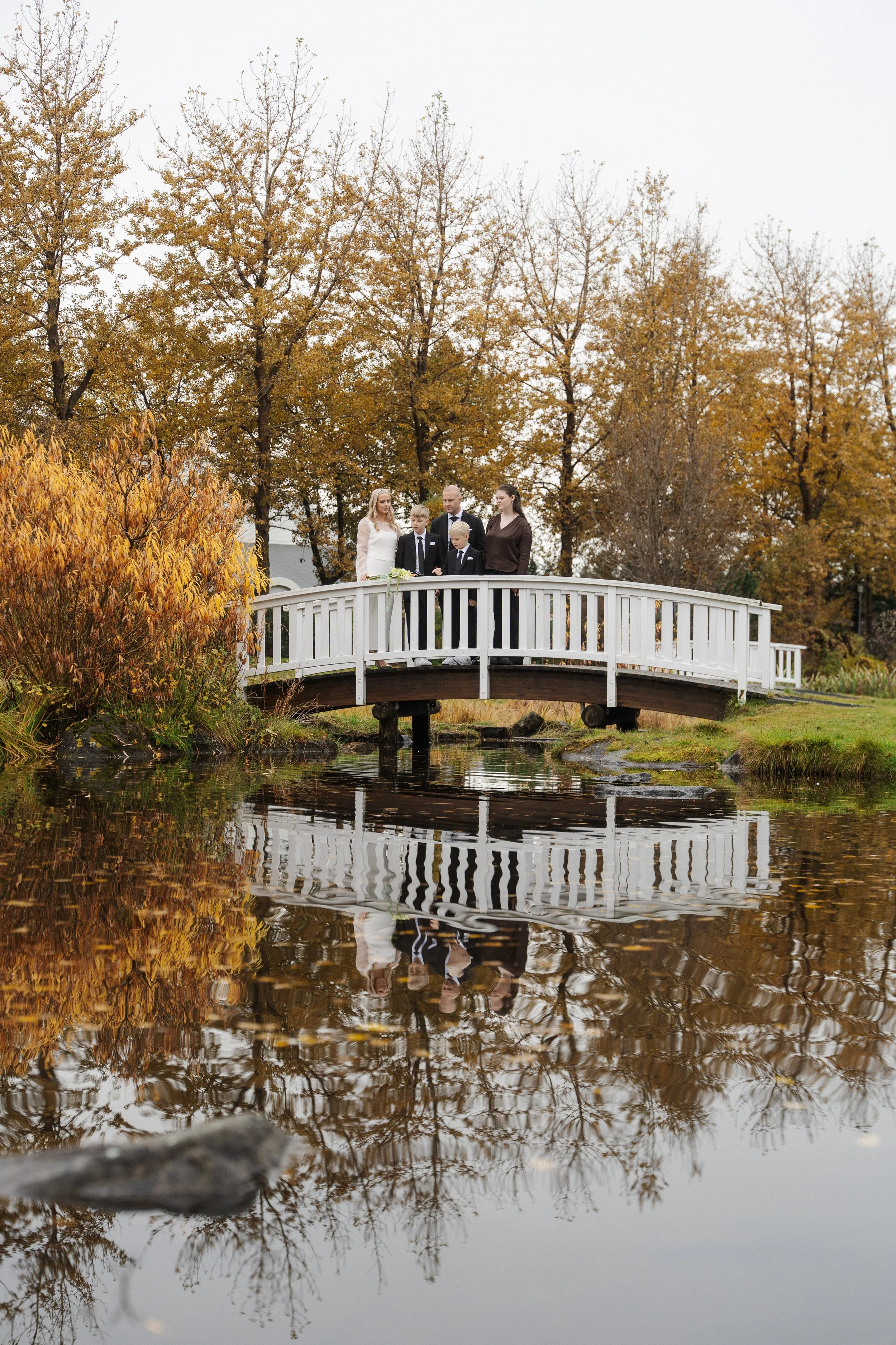 Group of five people standing on a small white footbridge over a reflective pond, surrounded by autumn trees.