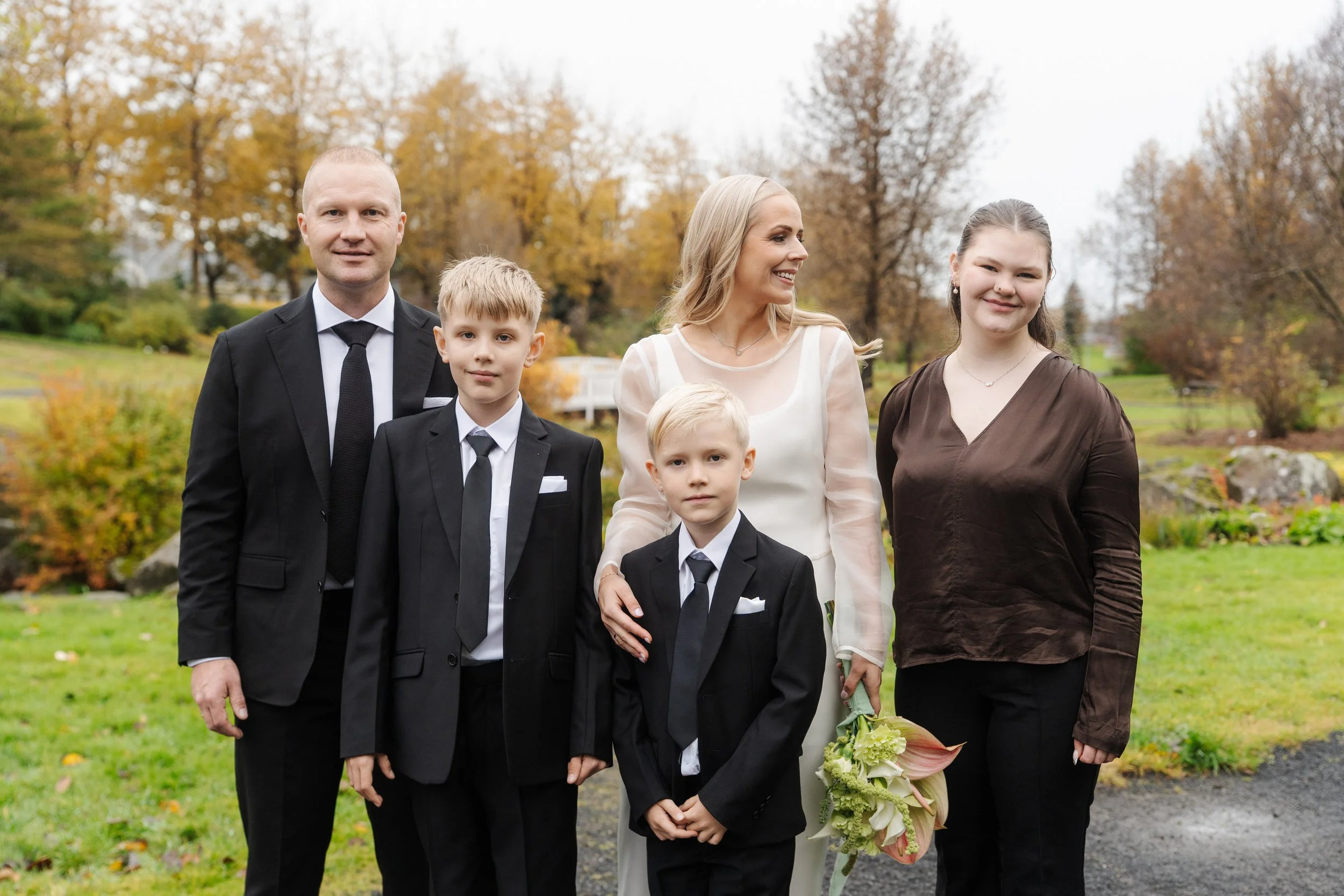 A family of five standing outdoors in a park with autumn trees in the background. The mother is holding a bouquet of flowers, and the children are dressed in formal attire with suits and ties.