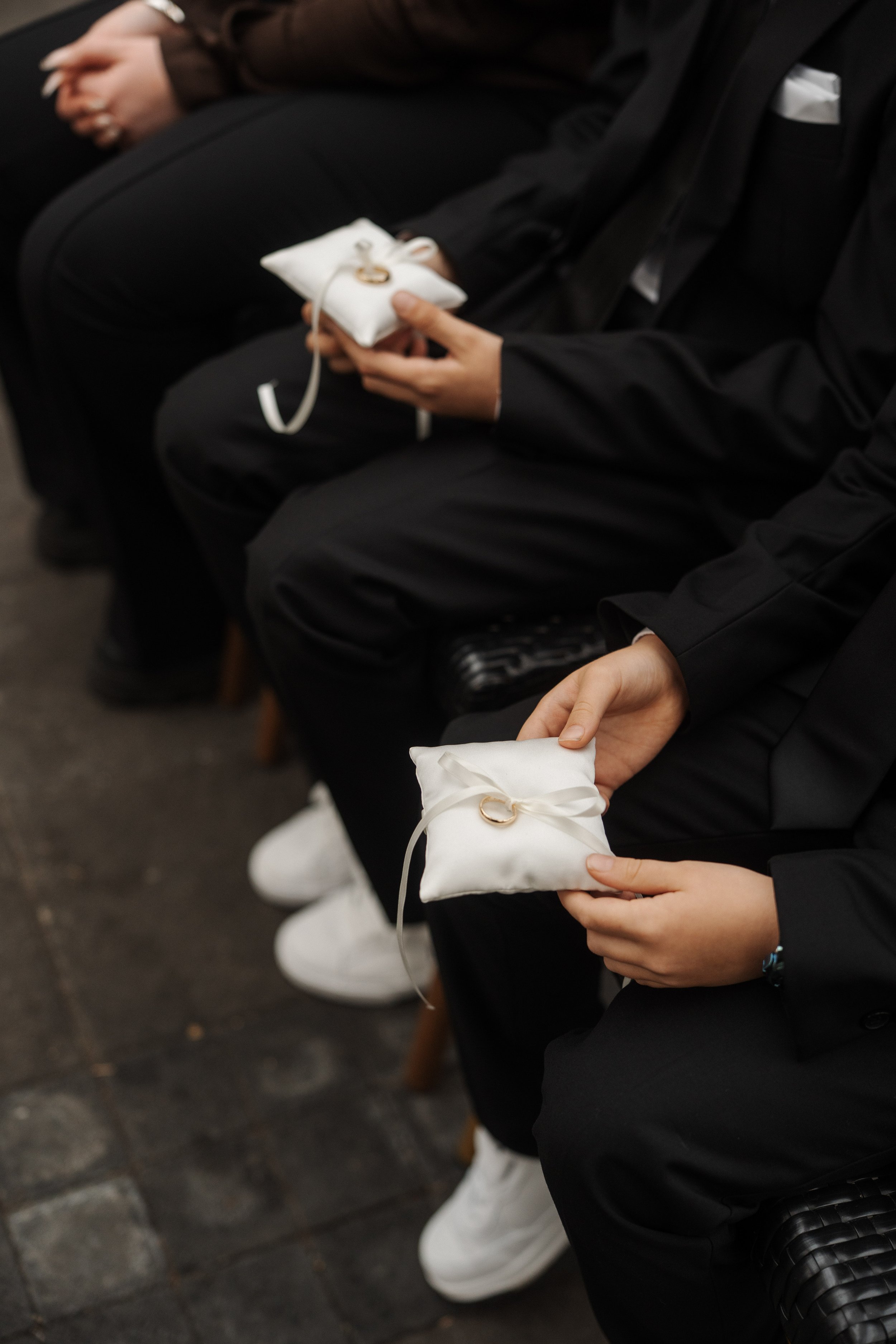 People dressed in black formal attire sitting on benches, each person holding a white ring pillow with a gold wedding ring.