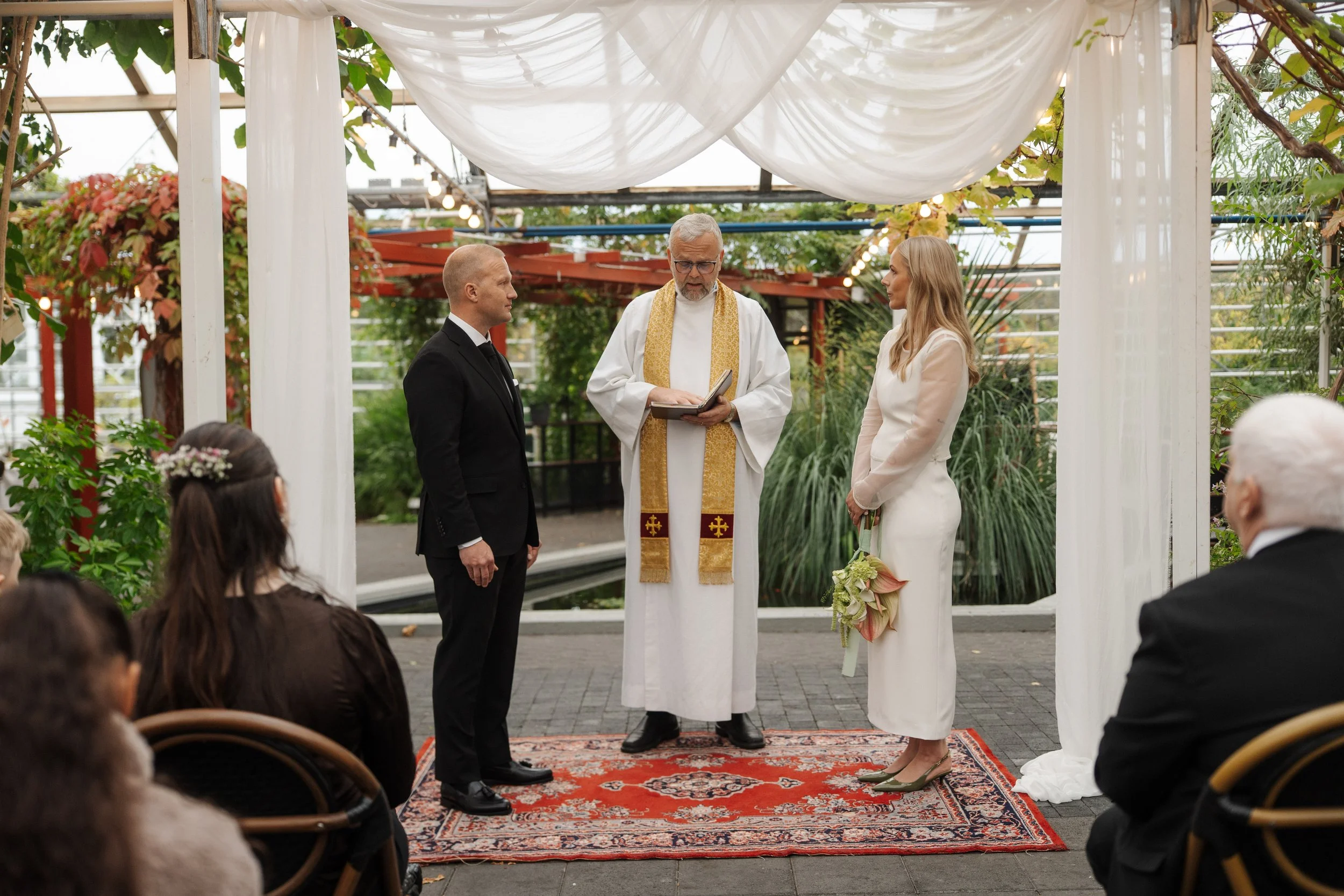 A couple getting married during an outdoor wedding ceremony, officiated by a priest in a white robe with a gold stole, standing on a patterned red rug under a draped white canopy with greenery around.