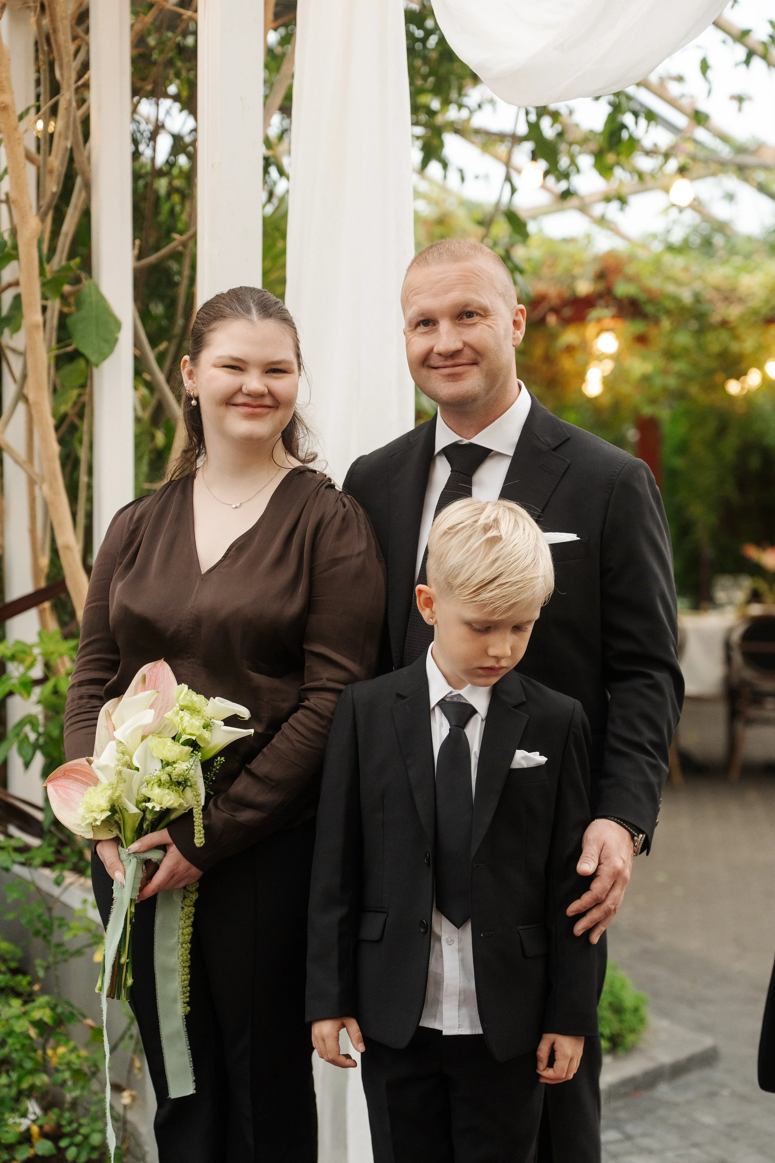 A family of three dressed in formal attire at a wedding or special event. The woman is holding a bouquet of flowers, standing next to a man and a young boy, all outdoors with decorative white drapes and green foliage around.