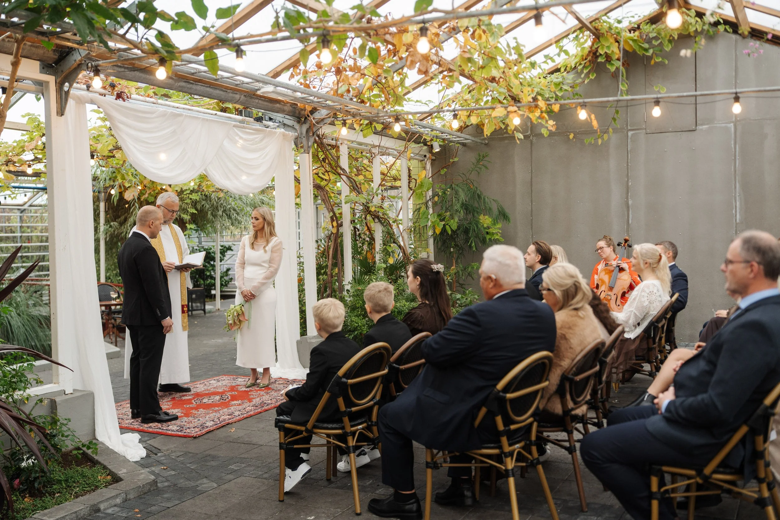 A wedding ceremony taking place in a greenhouse or garden setting with an audience, officiant, bride, and groom under a white canopy with string lights and greenery.