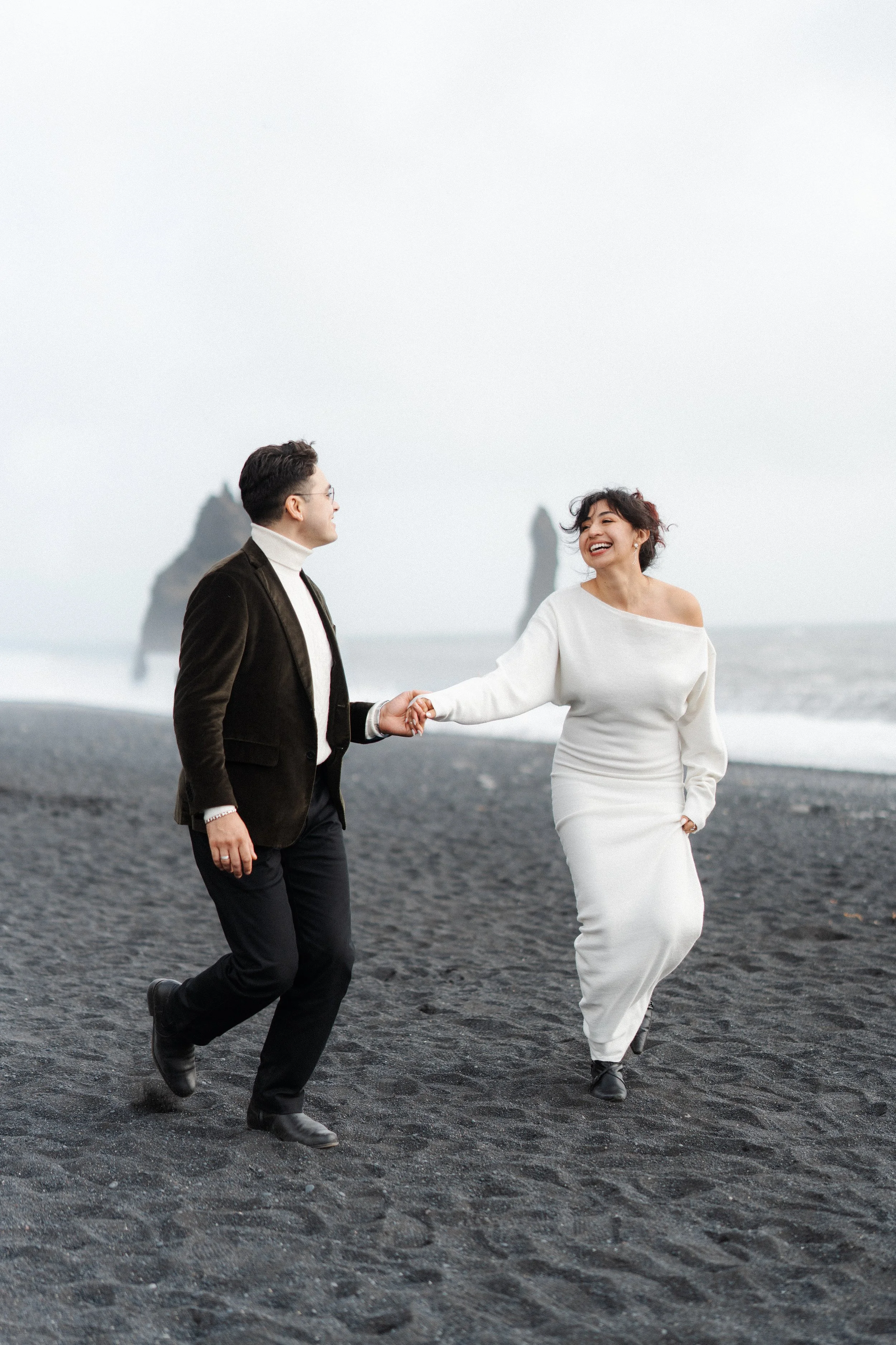 A couple holding hands and smiling while walking on a black sand beach with rock formations in the background.