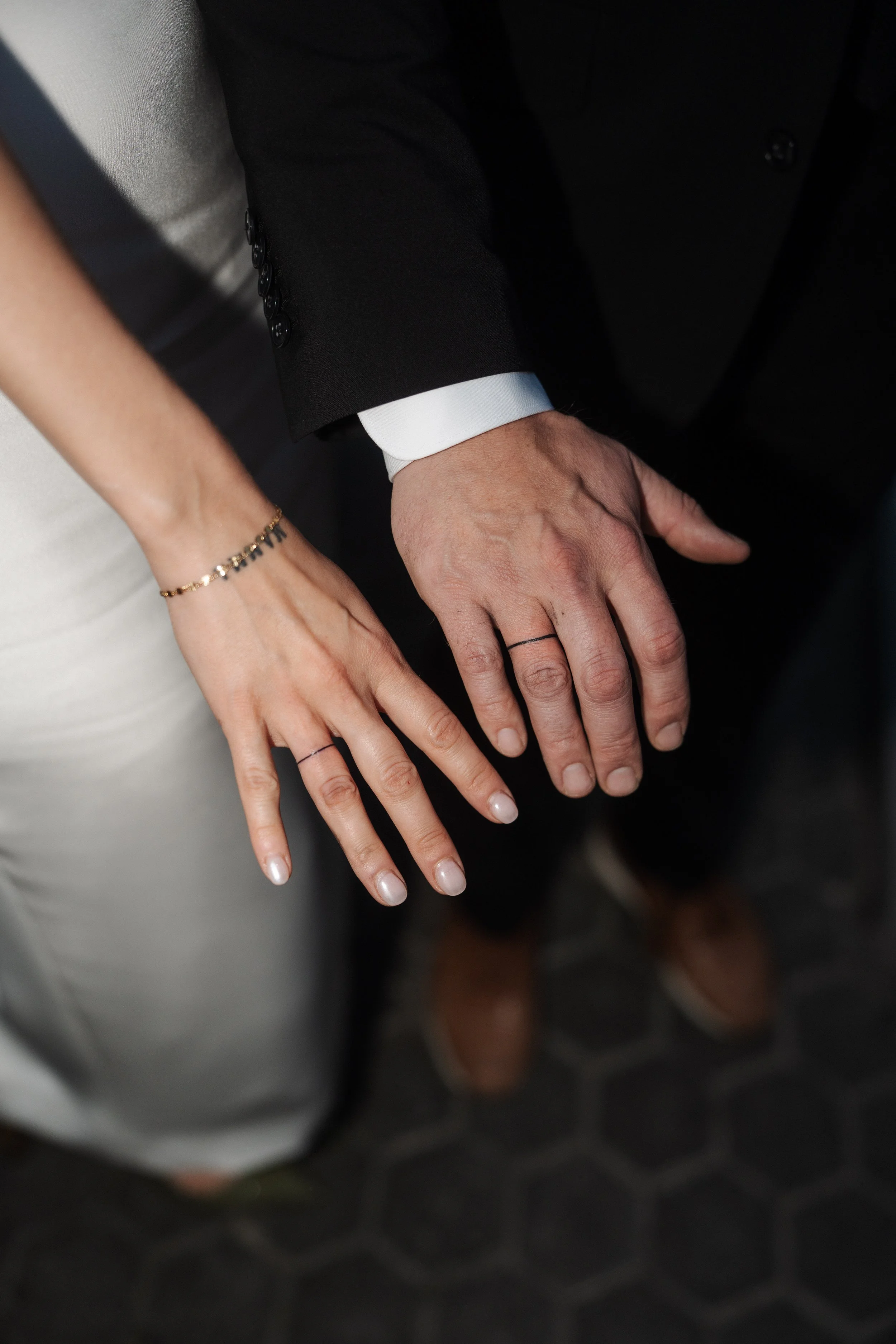 Close-up of a bride and groom's hands holding each other, showing wedding rings. The bride's hand has a gold bracelet and nails painted with light polish. The groom wears a black suit with a white shirt.