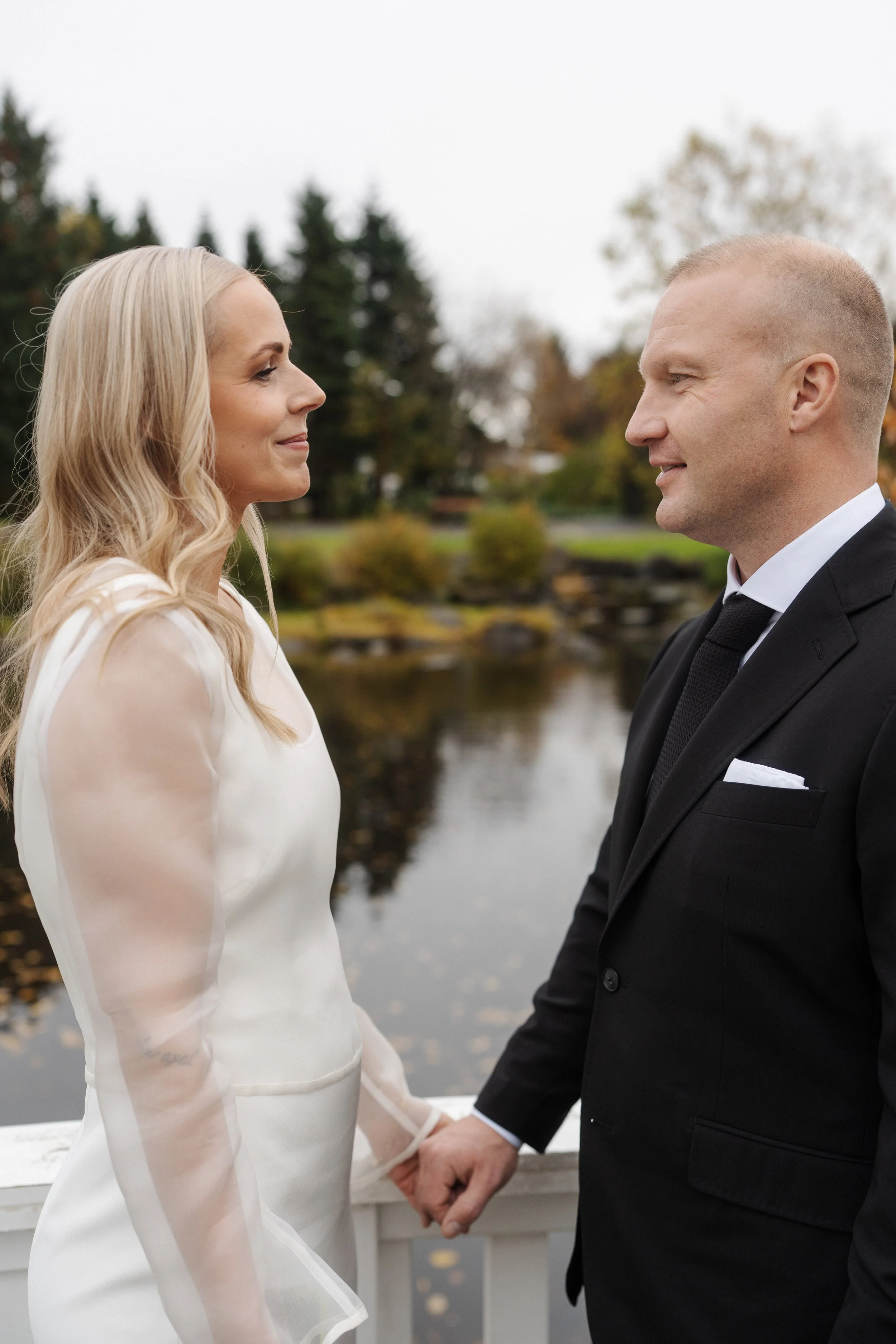 A couple in wedding attire holding hands and looking at each other outdoors near a lake with trees in the background.