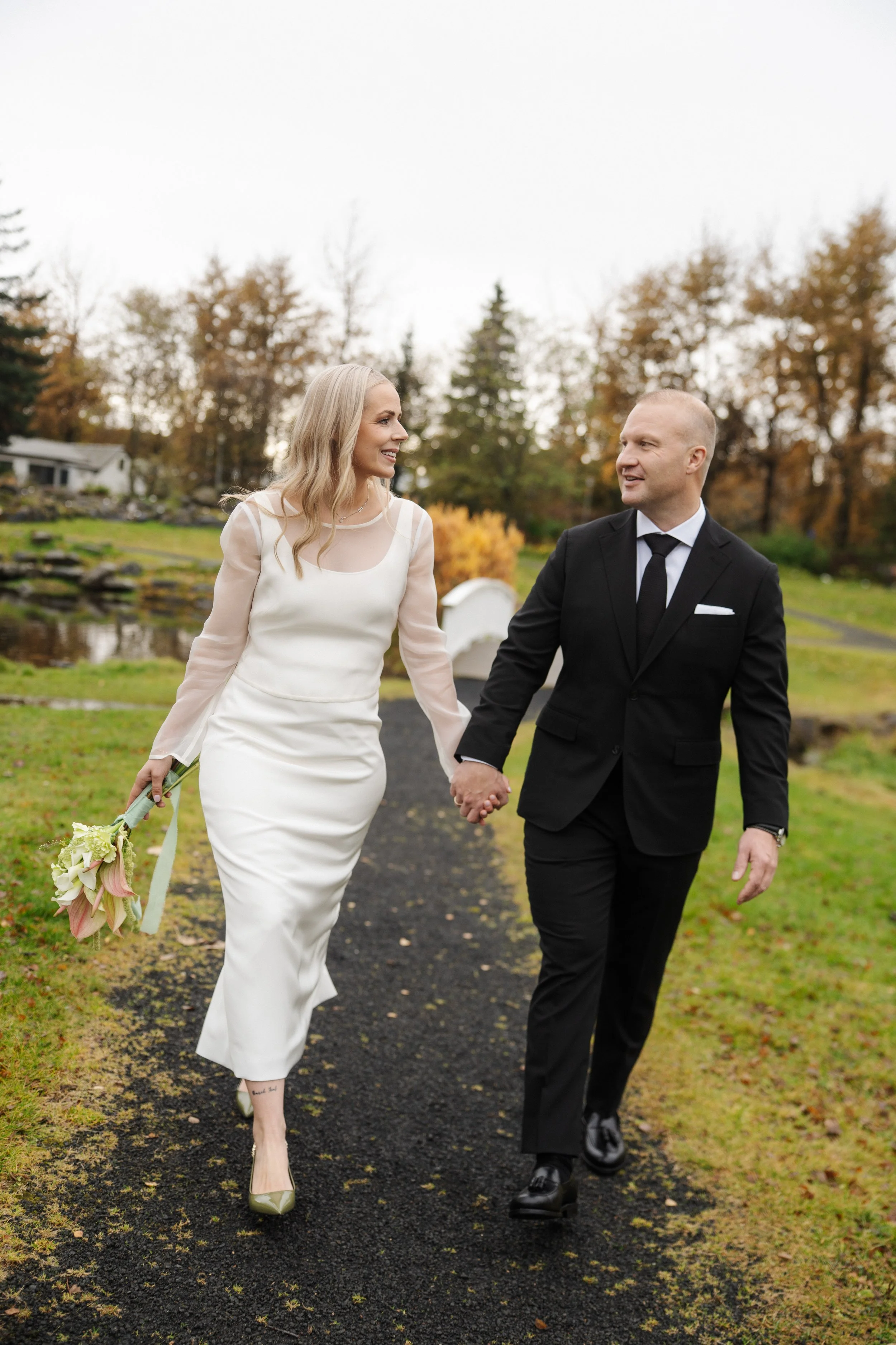 A newly married couple walking hand-in-hand outdoors on a cloudy day, with the bride in a white wedding dress holding a bouquet, and the groom in a black suit and tie, in a park with autumn trees.