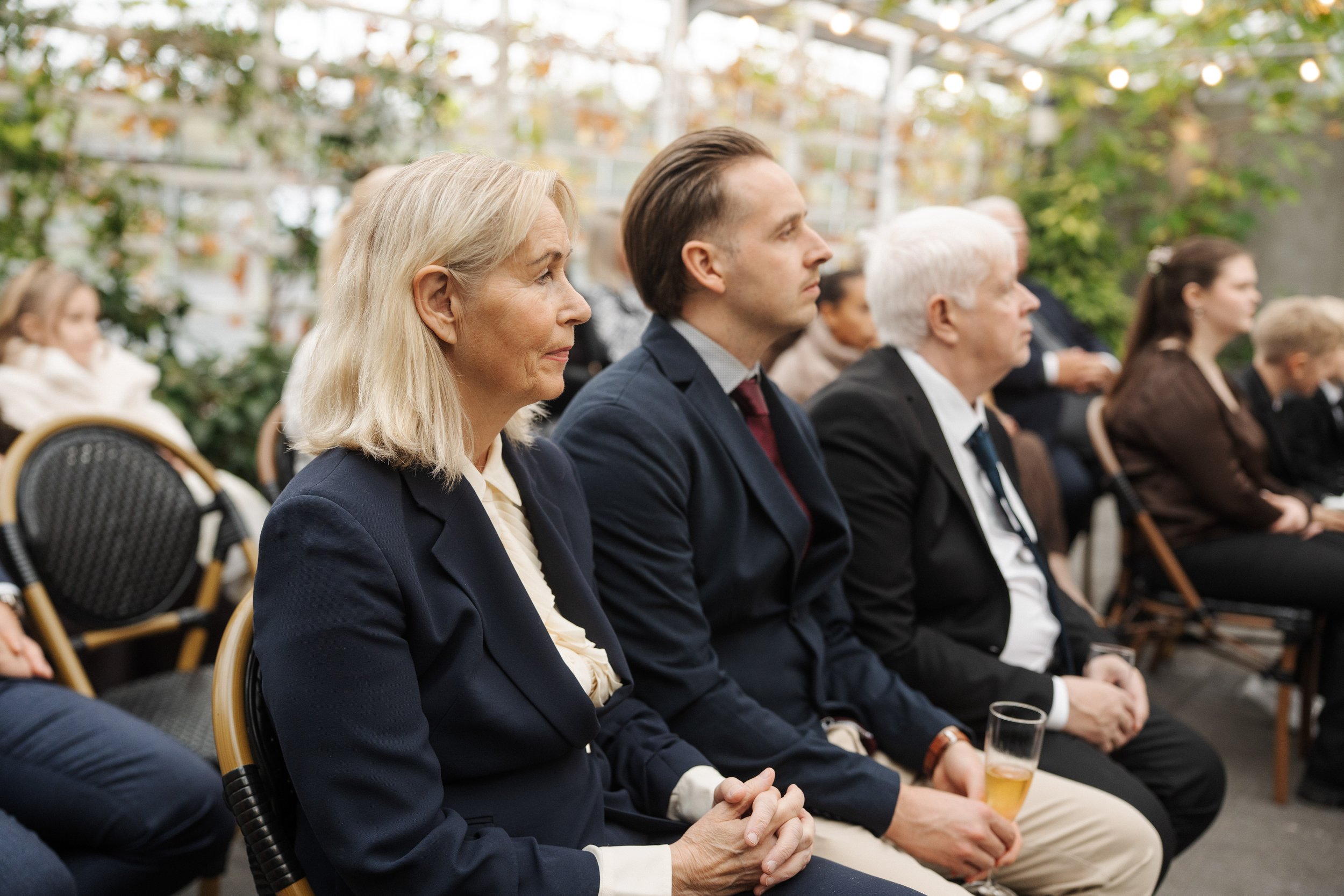 People sitting in an outdoor venue, attending an event or ceremony, dressed in formal attire, with some holding drinks, in a garden setting with trees and string lights in the background.