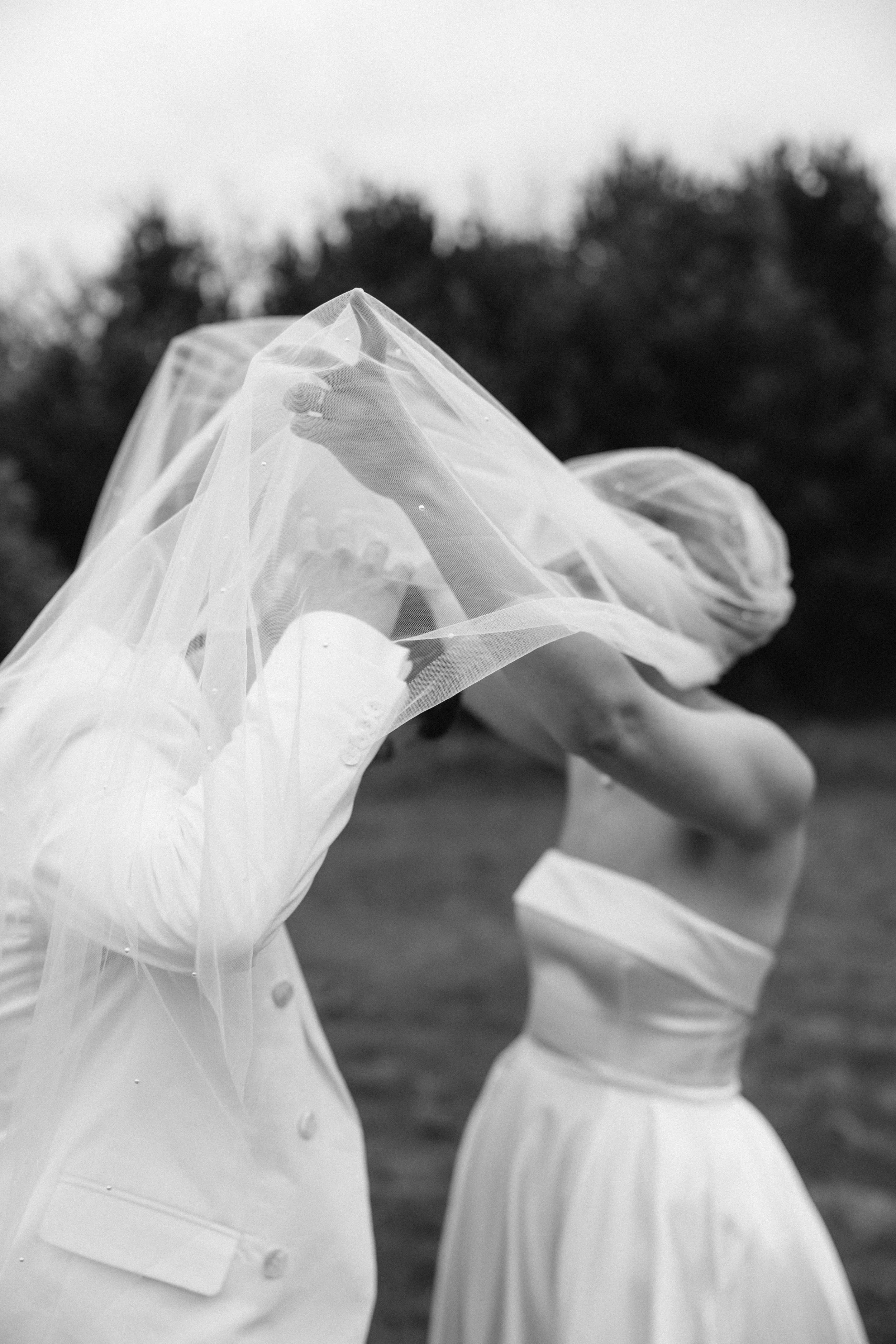 A person dressed as a bride and a person dressed as a groom cover themselves with a sheer veil outdoors at sunset.