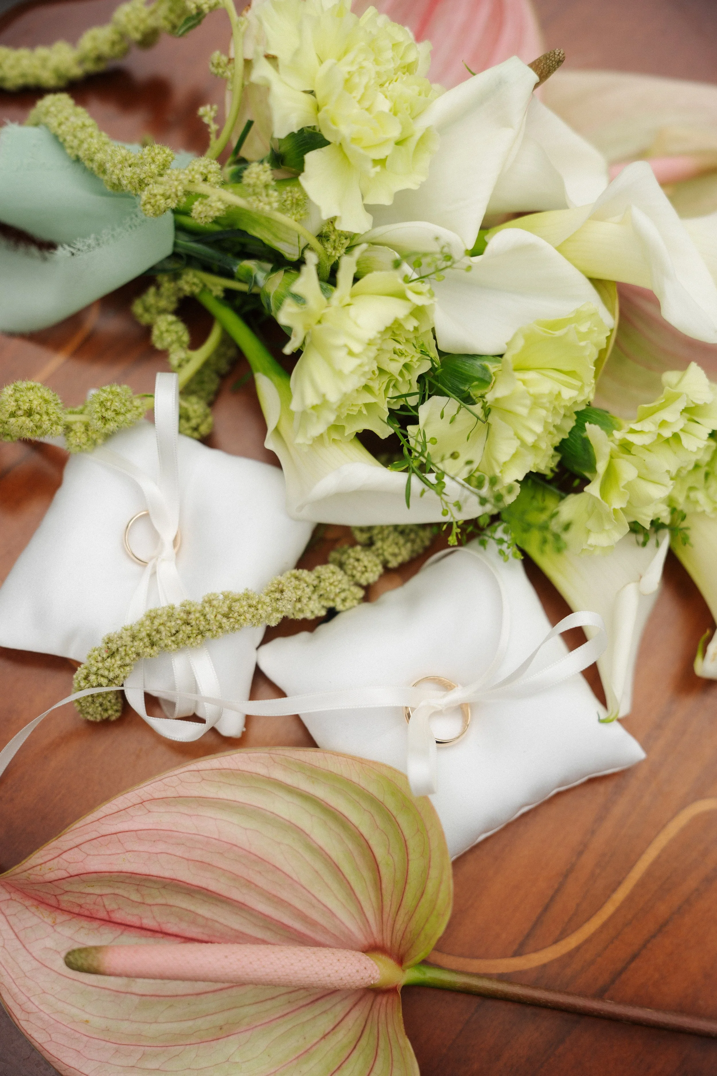 White wedding rings attached to white pillows with white ribbons, surrounded by a bouquet of cream and white flowers and a pink and green anthurium flower.