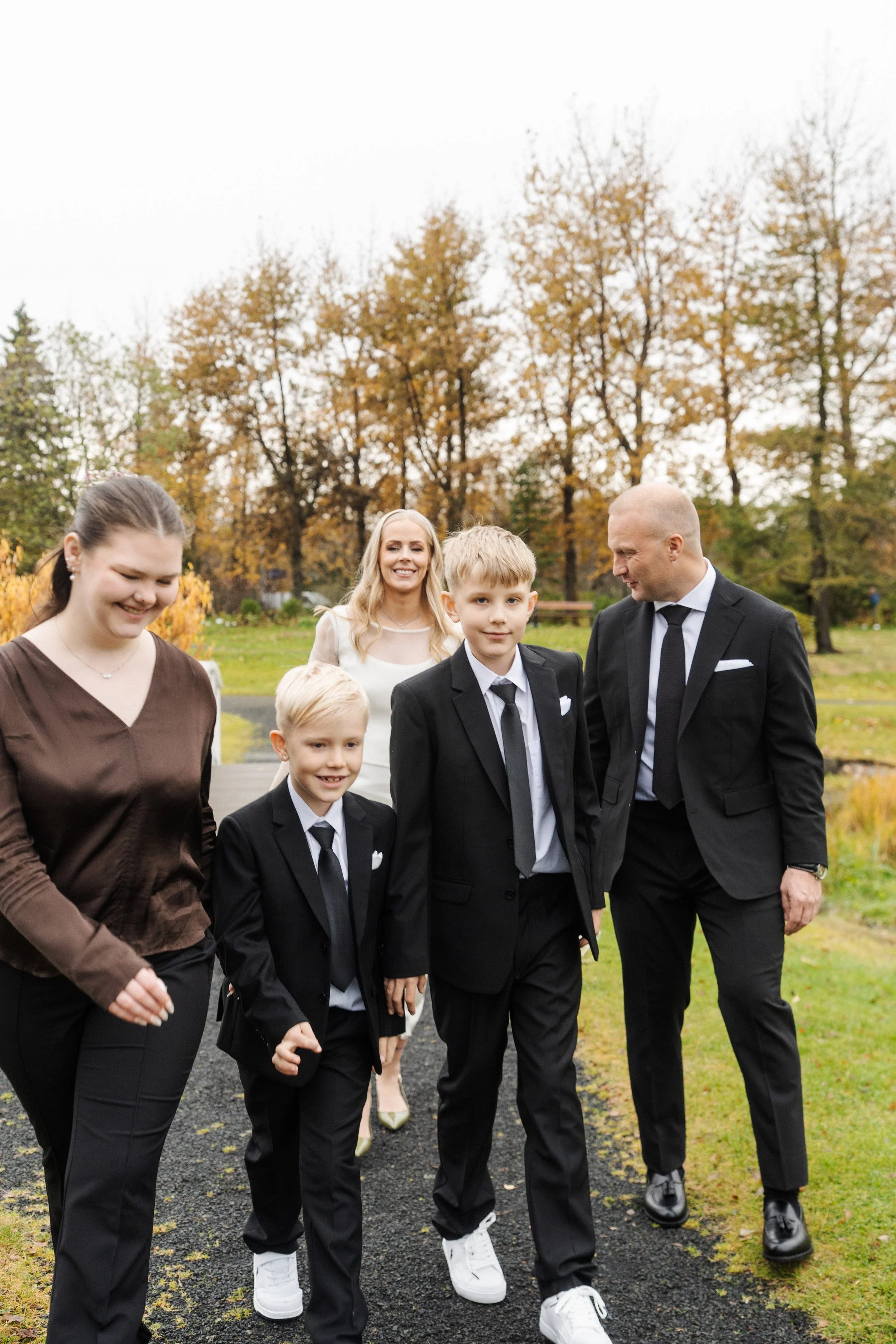 A family walking outdoors in a park during autumn, dressed in formal attire, with trees and fall foliage in the background.