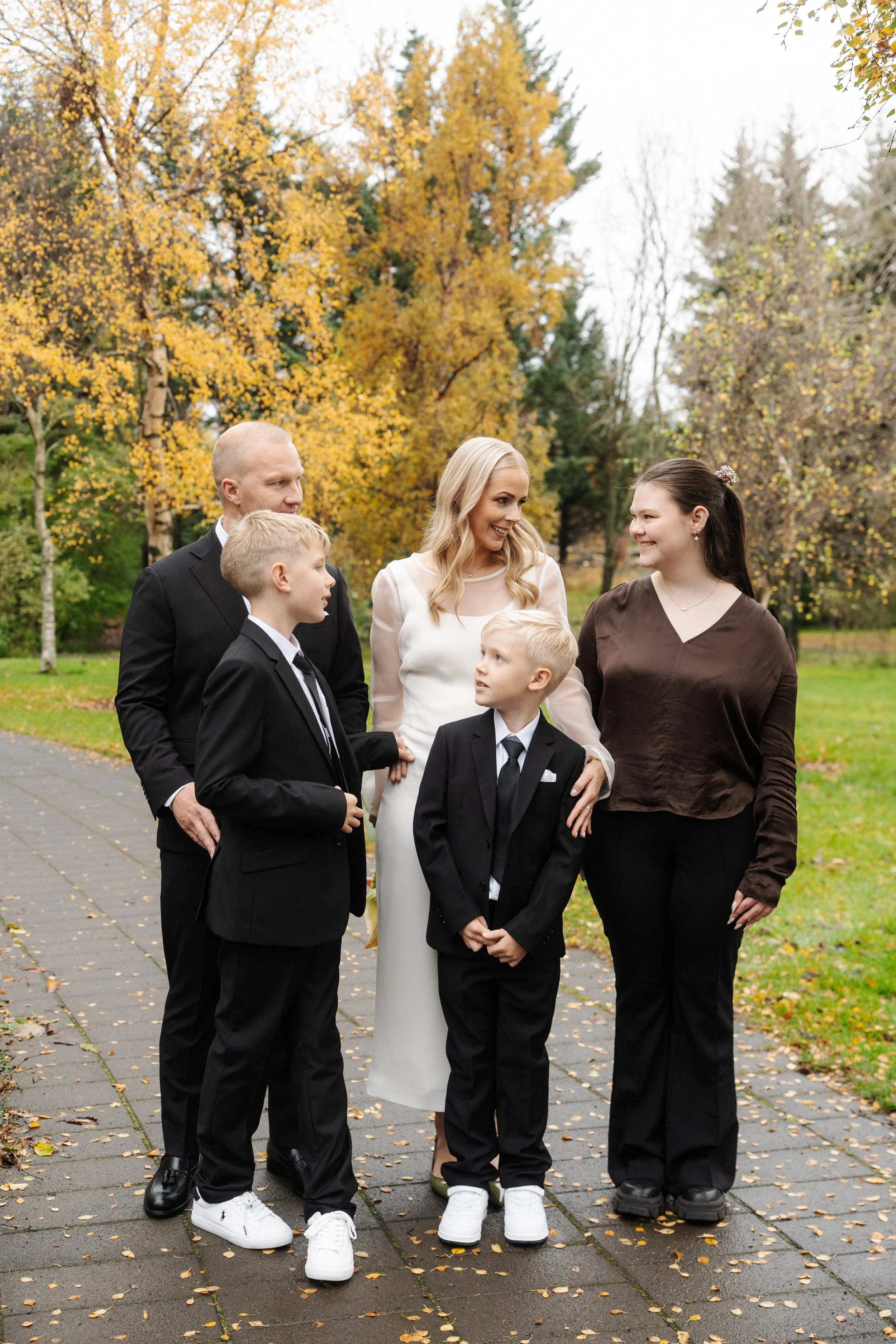A group of six people, including two women, one man, and three boys, standing outside on a sidewalk in fall with colorful autumn trees in the background, dressed in formal and semi-formal attire, engaging in conversation.