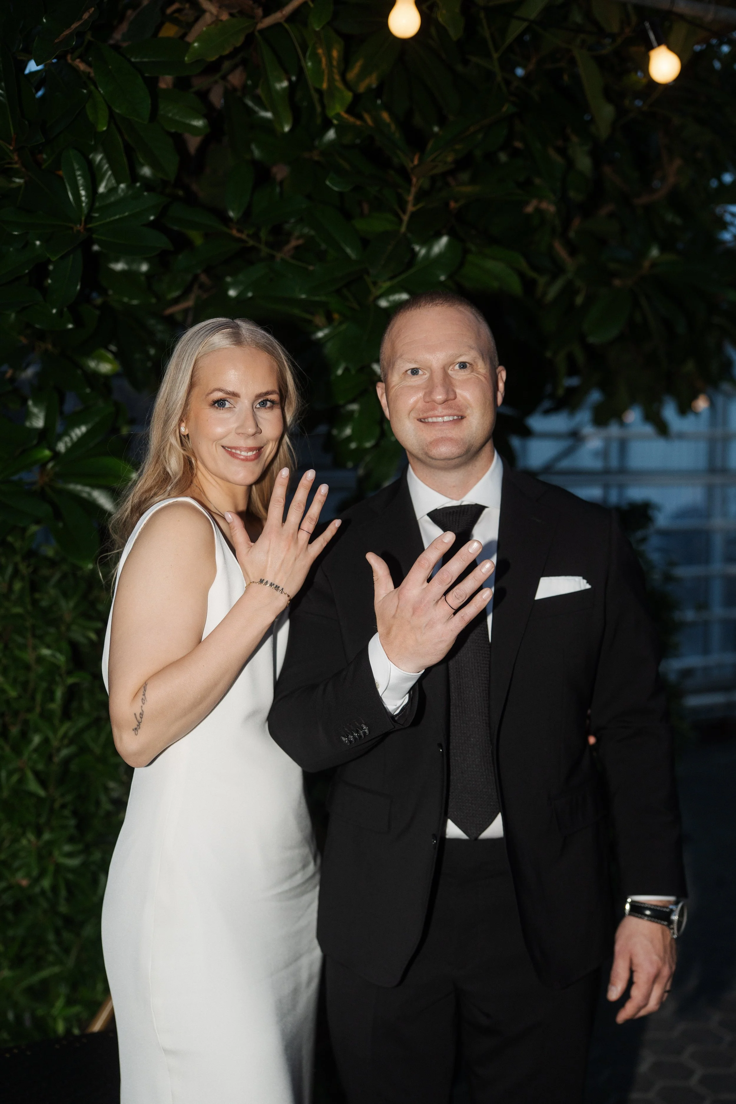 A newly married couple displaying their wedding rings and smiling at camera, standing in front of leafy green background with hanging lights.